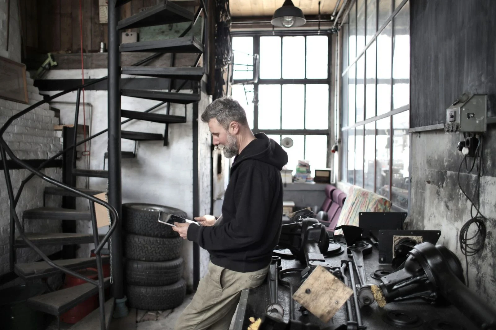 A bearded mechanic in work clothes uses a tablet beside a workbench in a workshop.