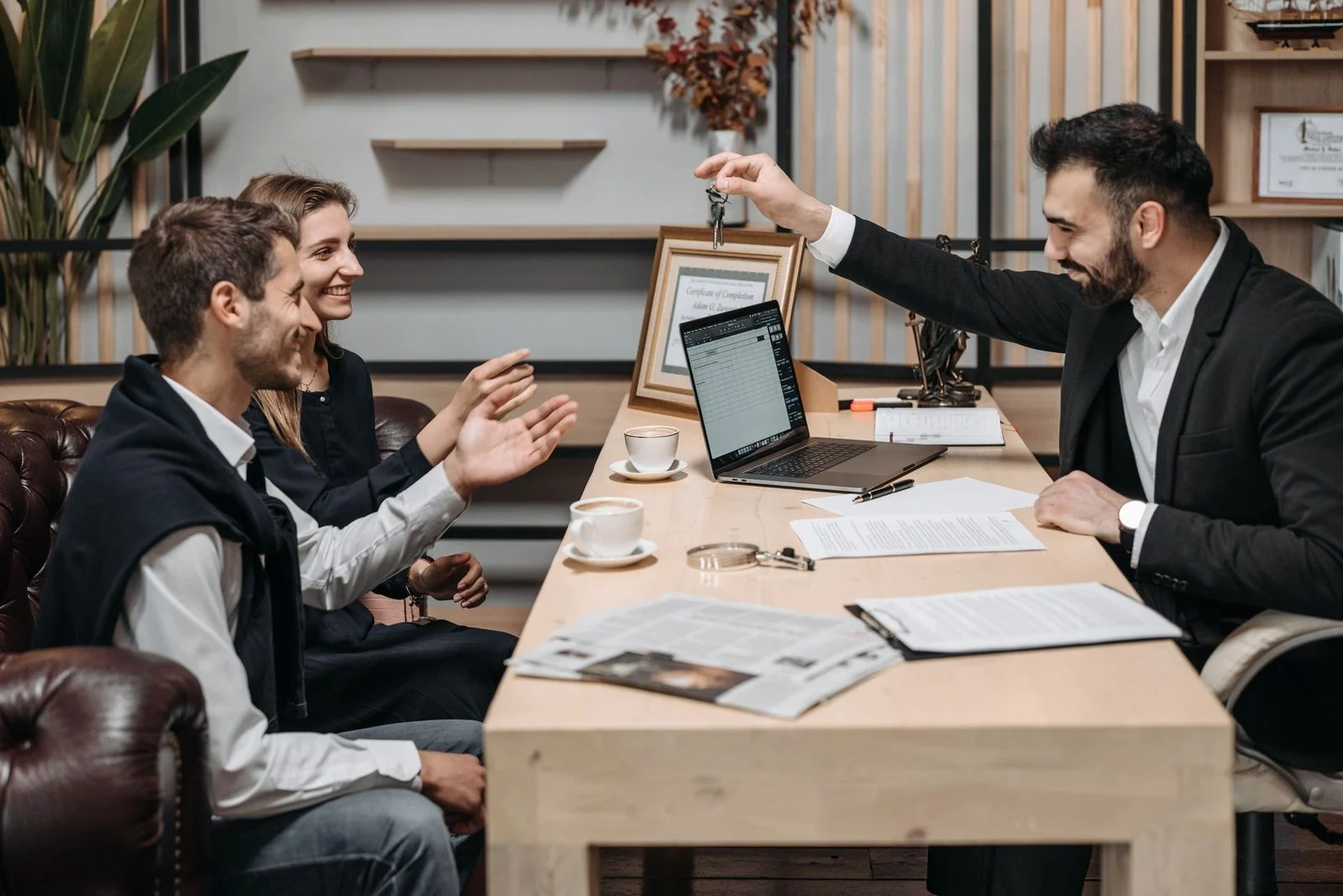 A business professional hands keys to smiling clients across an office desk during a consultation.