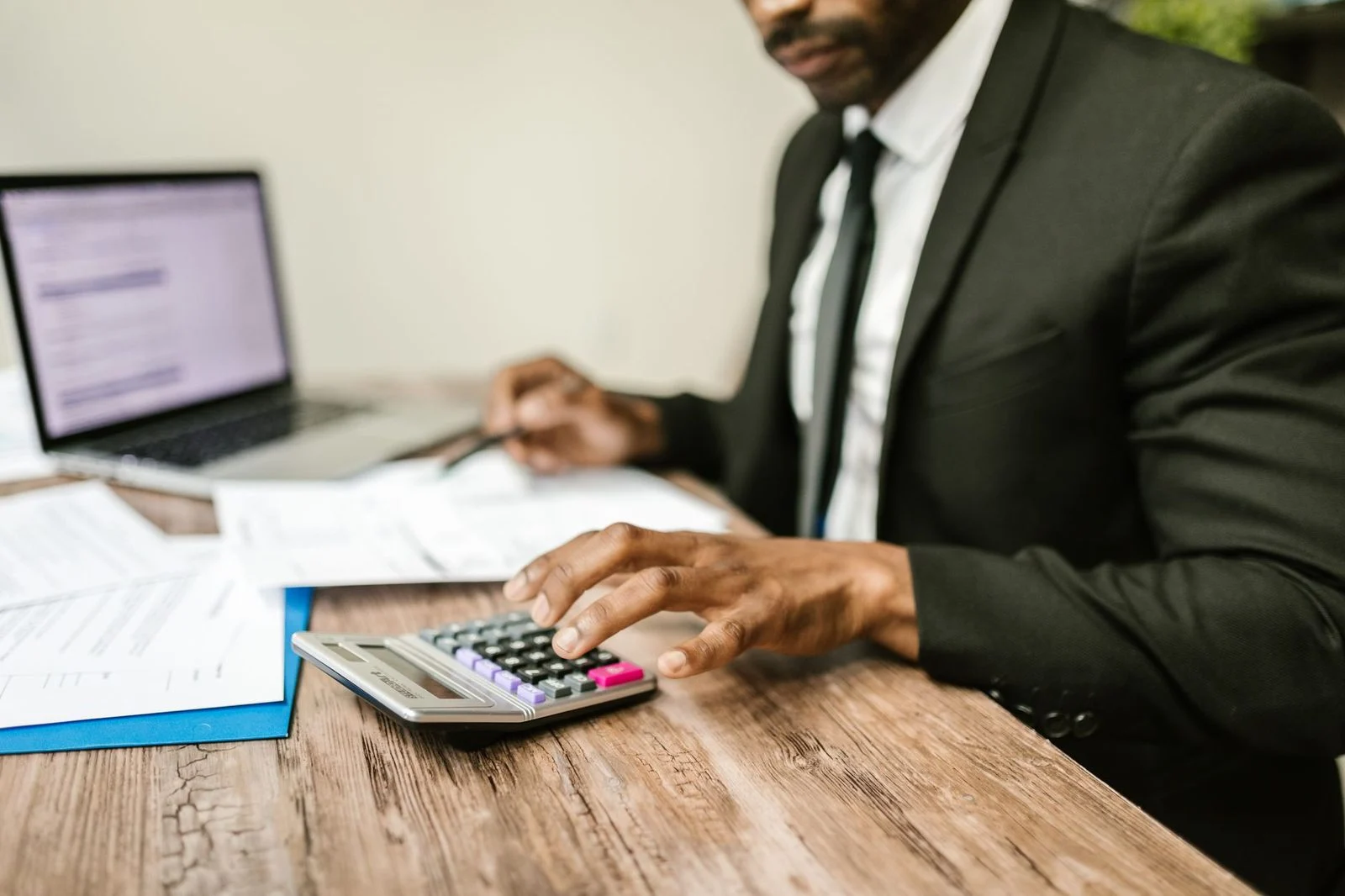 Hombre de negocios con traje negro usando una calculadora en un escritorio junto a documentos financieros y una laptop.