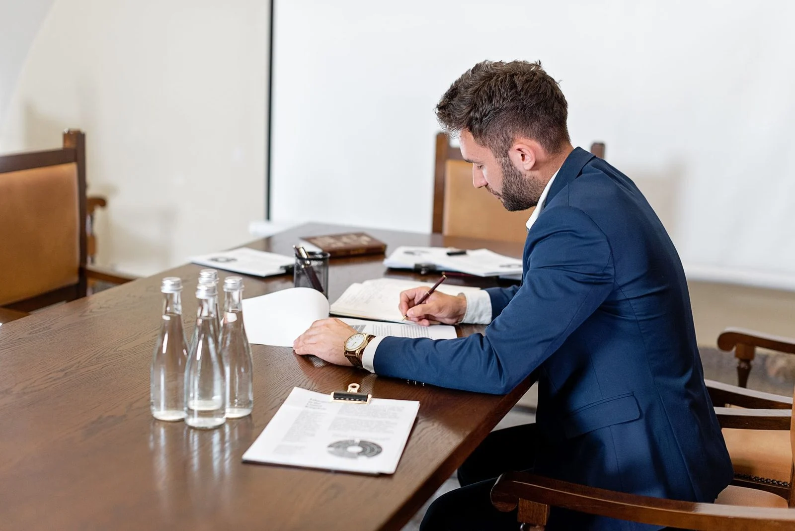 A businessman in a blue suit signs a contract at an office desk beside paperwork and water bottles.