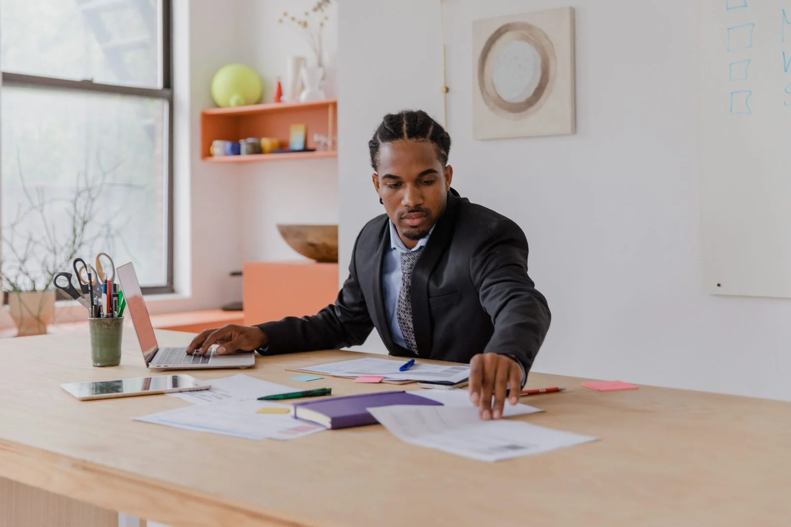 A businessman sits at a modern desk in an office, reviewing policy documents and a financial projection spreadsheet beside a framed family photo.