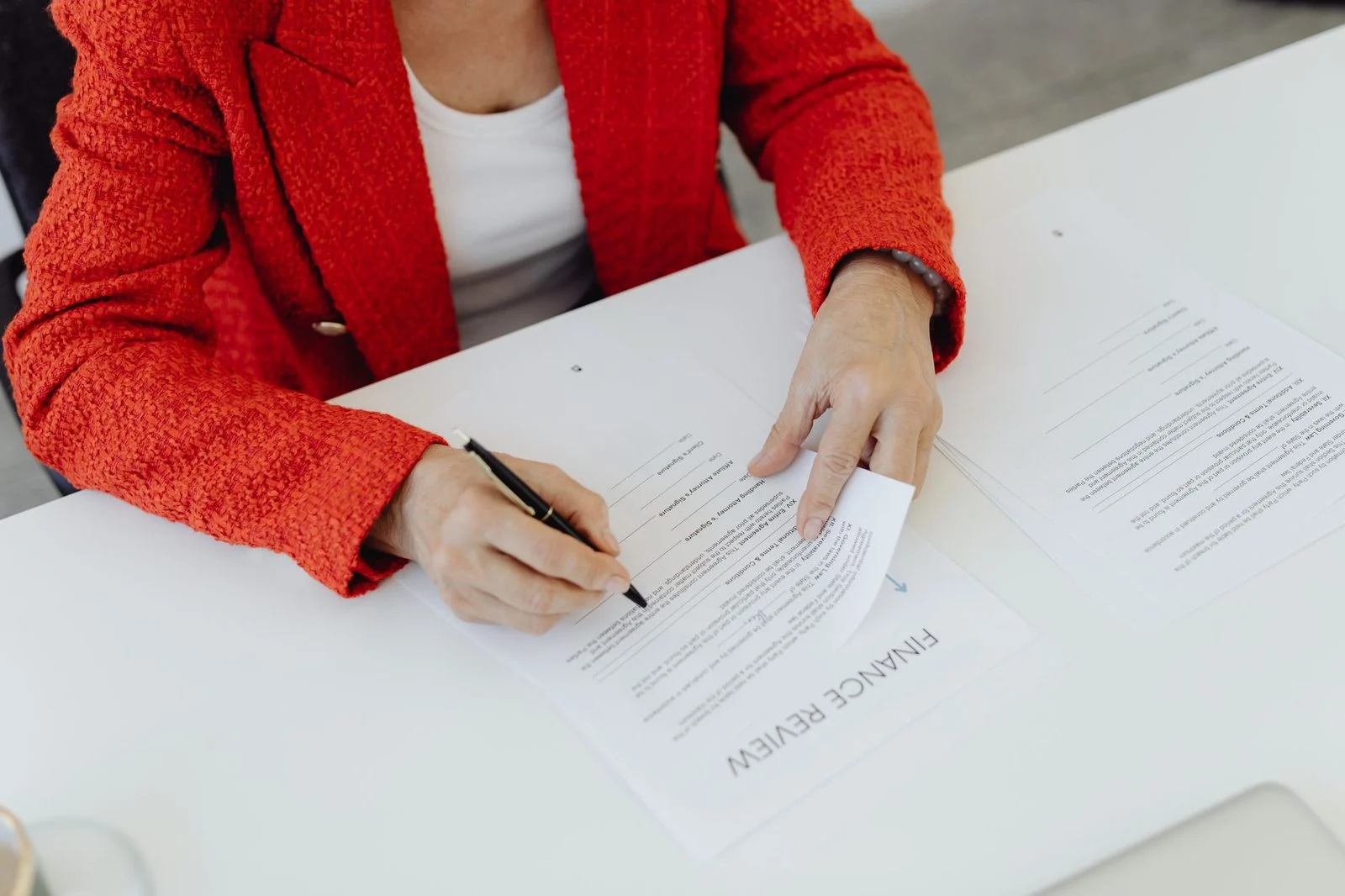 A businesswoman in a red blazer signs financial documents at an office desk.