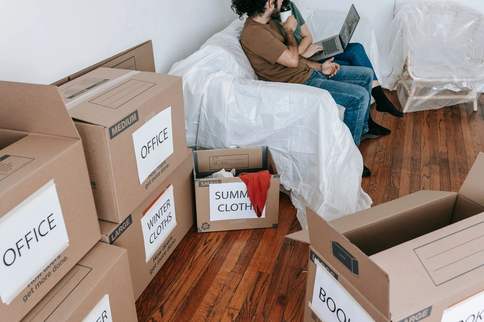 Una pareja sentada en un sofá rodeada de cajas de mudanza etiquetadas en una habitación luminosa.