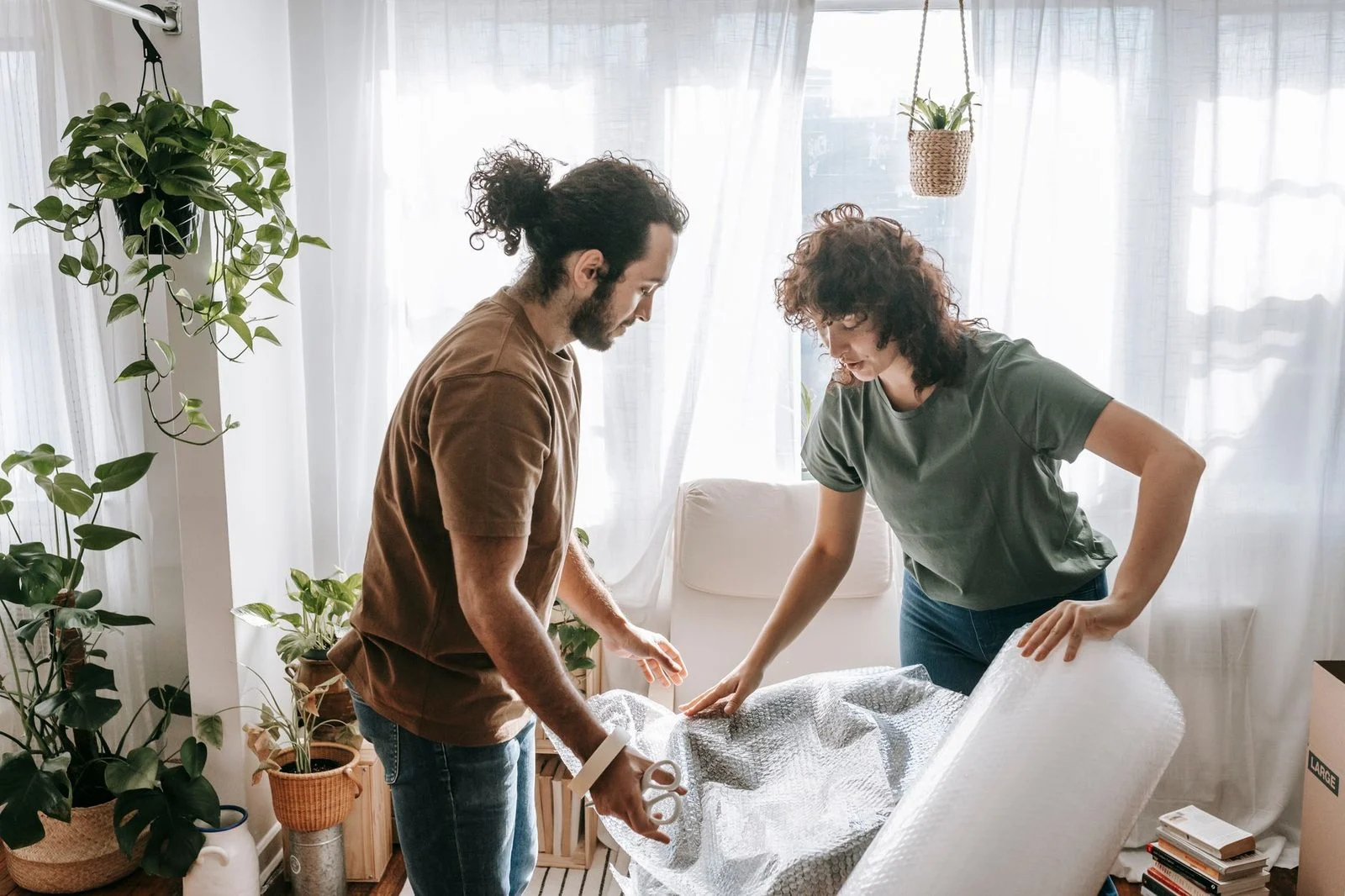 A couple wraps furniture in a plant-filled living room while preparing for a move.