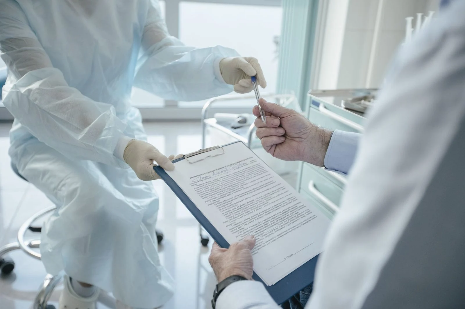 A doctor hands a clipboard to a patient to sign paperwork in a bright clinic exam room.