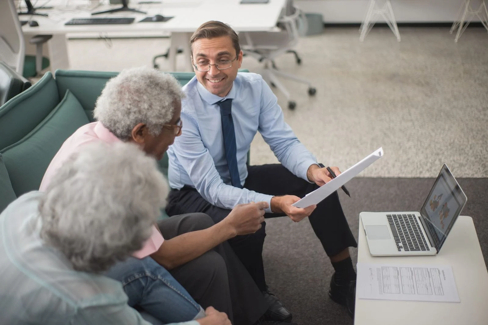 A financial consultant reviews documents and a laptop with two senior clients in a bright, modern office.