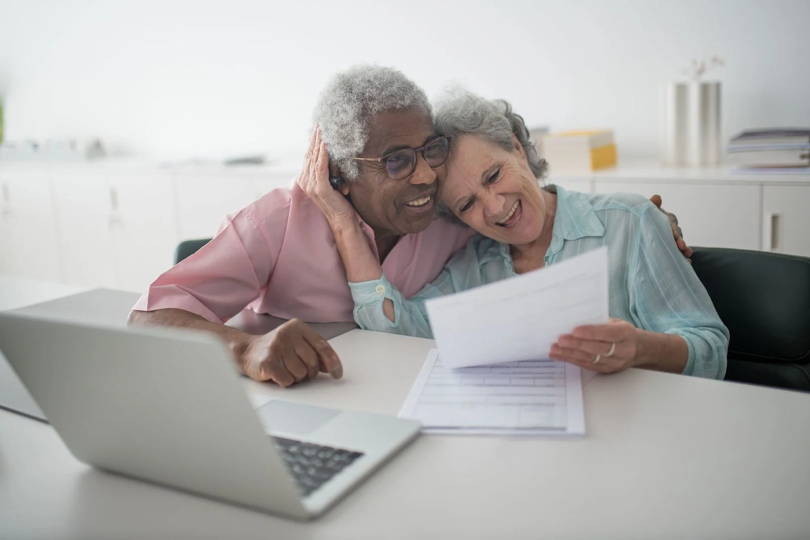 Una pareja de adultos mayores sonriente revisa documentos juntos en un escritorio junto a una laptop.