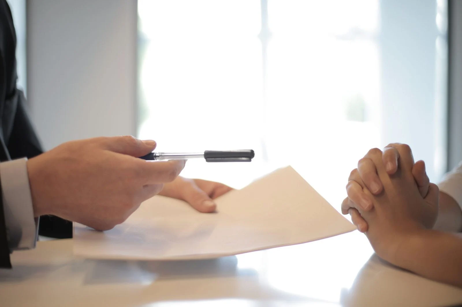 A man in a suit hands a pen and paper across a table to a woman with clenched hands during a business meeting.