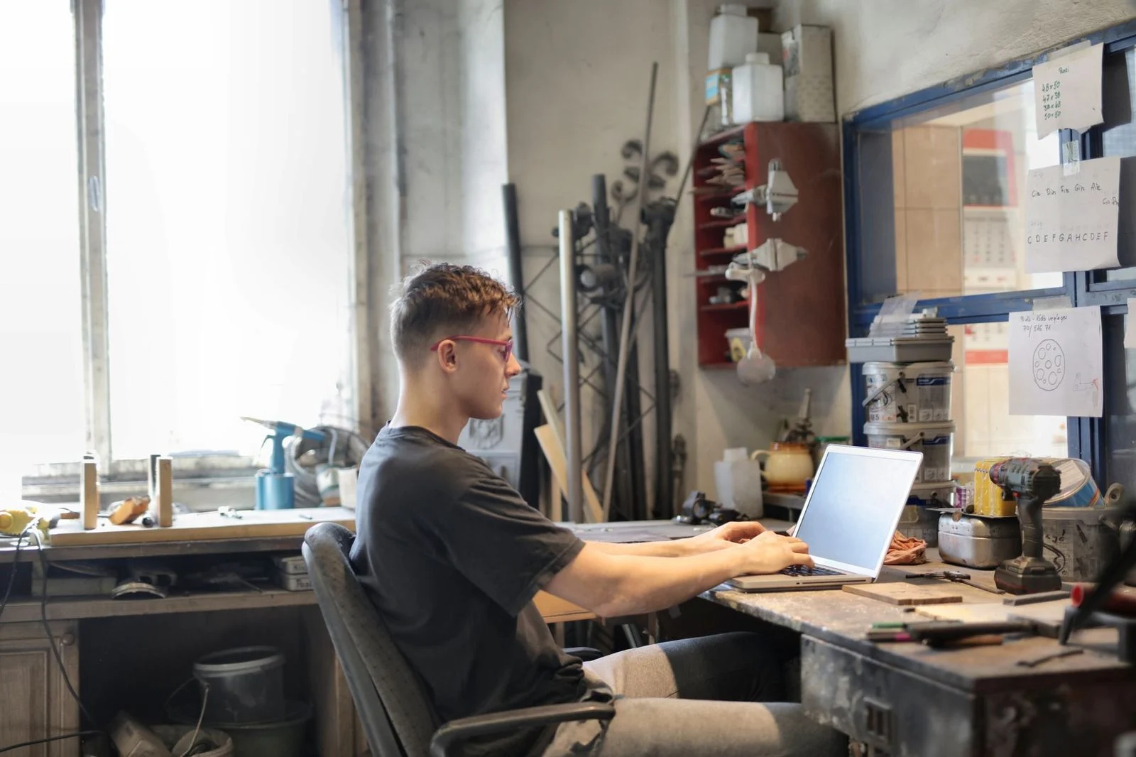 A man in eyeglasses and casual clothes sits at a table in a workshop, working on a laptop.
