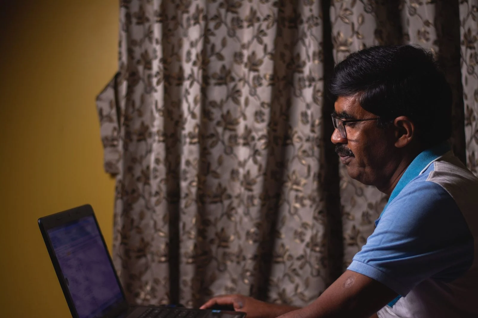 A man wearing glasses works on a laptop at home.