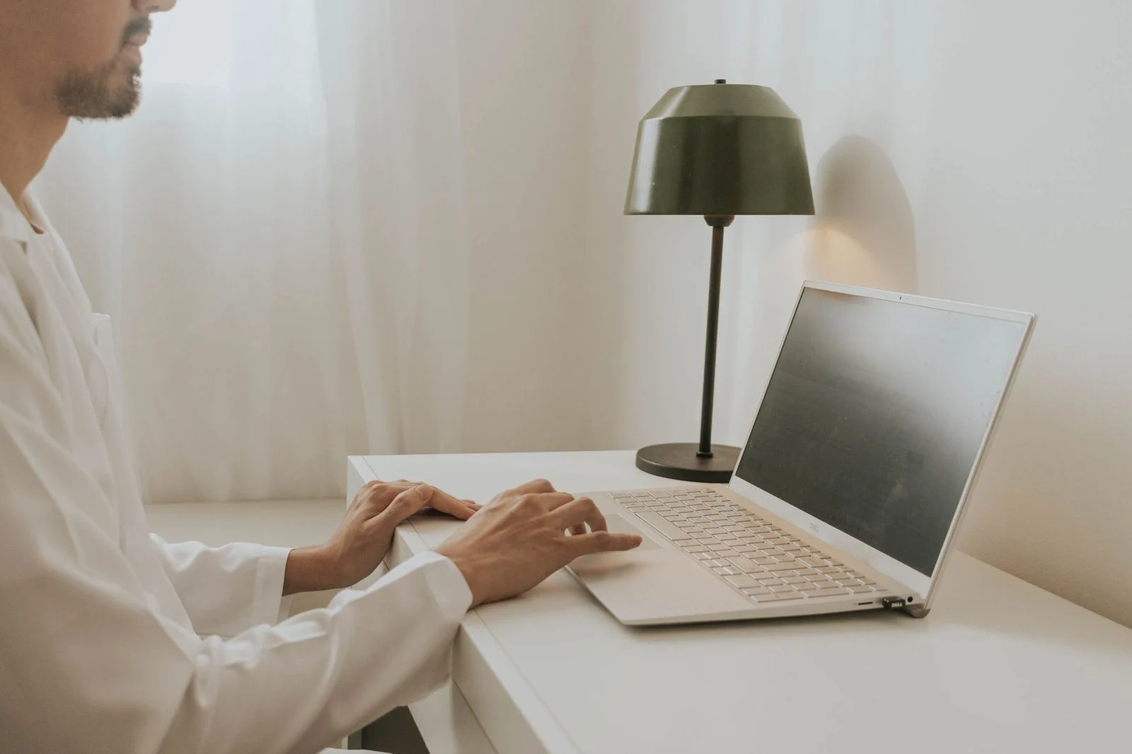 A man works on a laptop at a minimalist desk beside a lamp in a home office.
