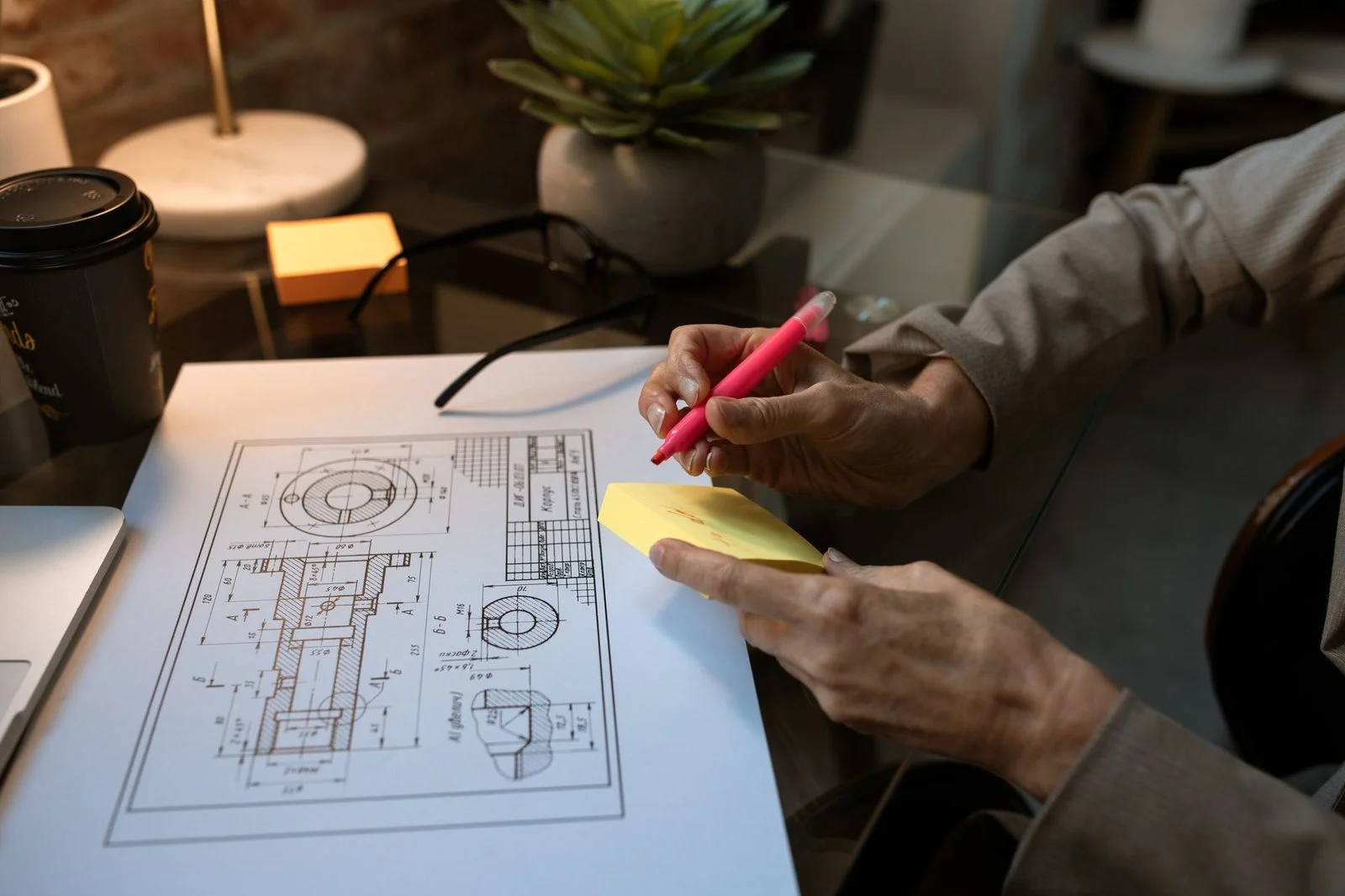 Una persona anota un plano técnico junto a una laptop y una taza de café en un espacio de trabajo moderno.