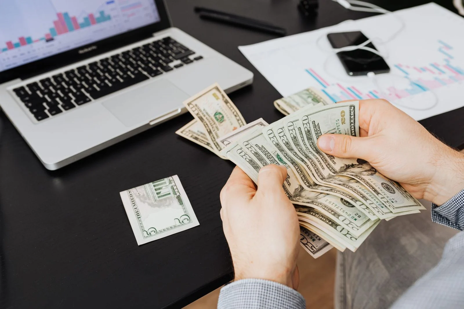 Una persona cuenta billetes de dólar en un escritorio junto a una laptop que muestra gráficos financieros.