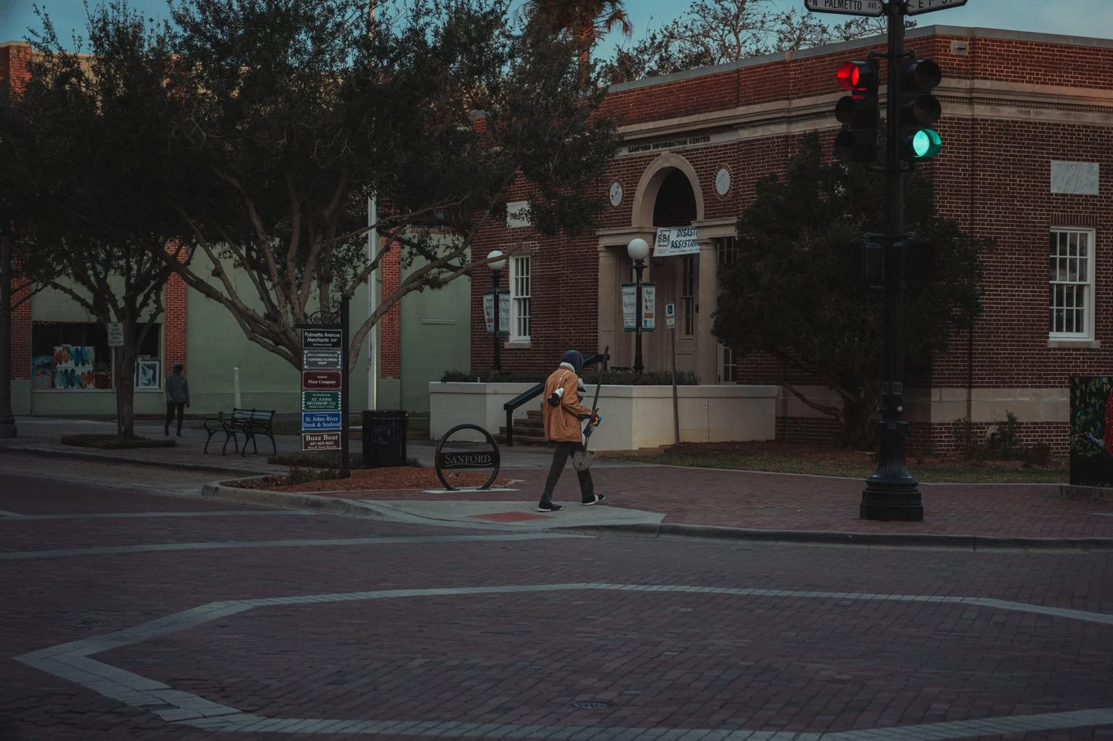 A person walks through a city street intersection at twilight, past brick buildings and trees.
