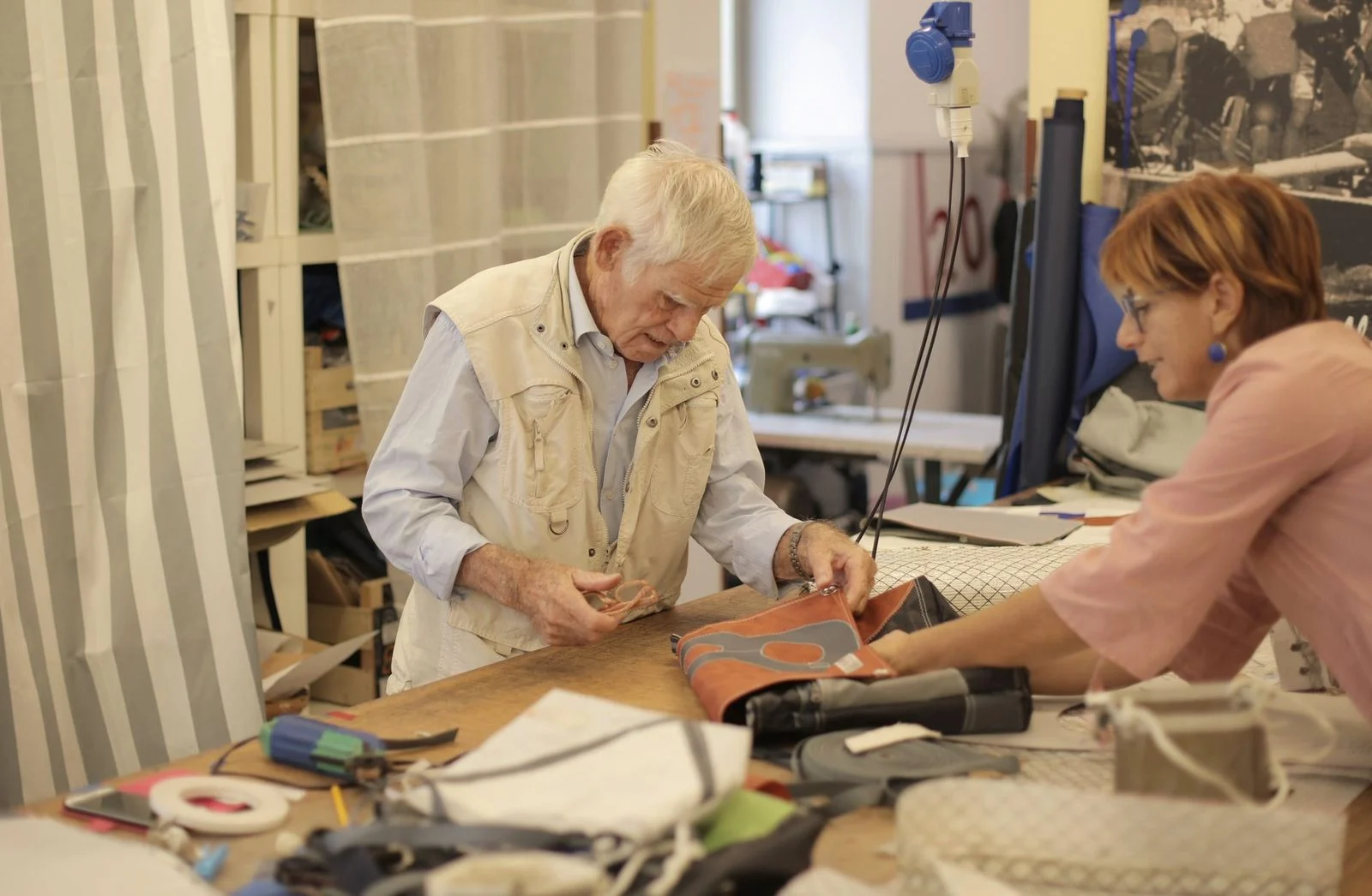 A senior male craftsman in casual clothes stands at a worktable shaping a leather accessory beside a female partner.