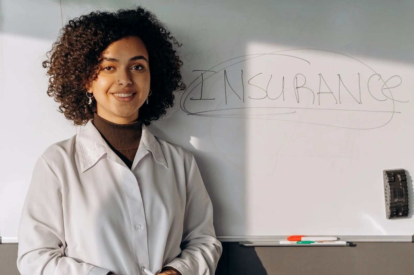 A smiling African American woman stands in an office in front of a whiteboard that reads "Insurance.