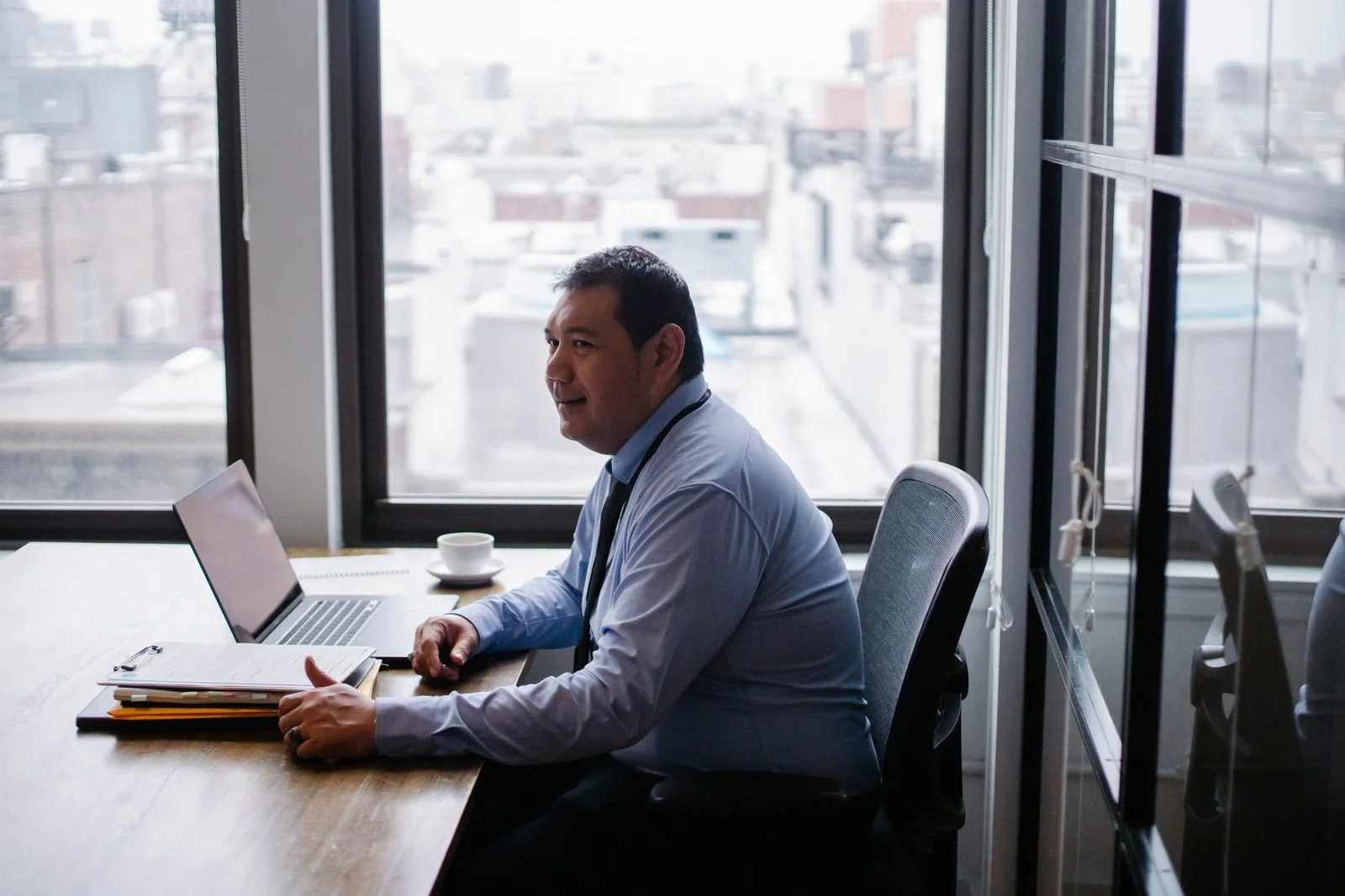 A smiling businessman sits in an armchair at a table, working on a laptop with documents spread out.