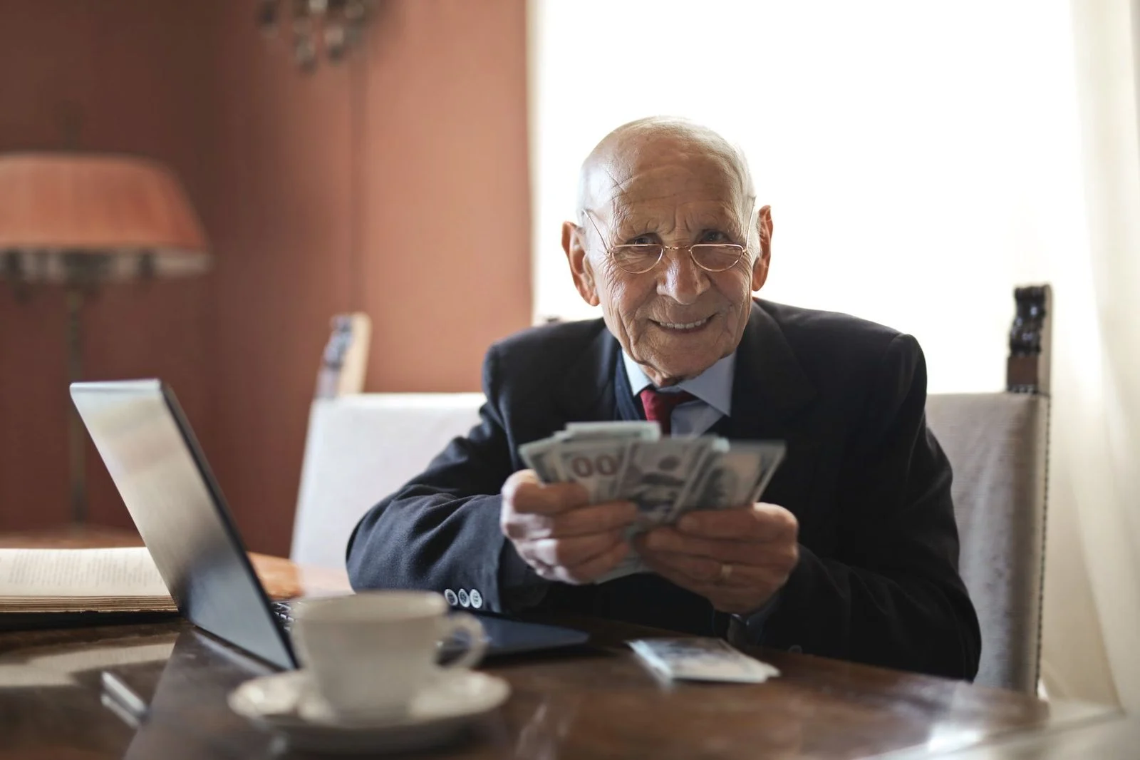 Hombre de negocios mayor y sonriente, con traje y gafas, sostiene billetes junto a una laptop sobre una mesa.
