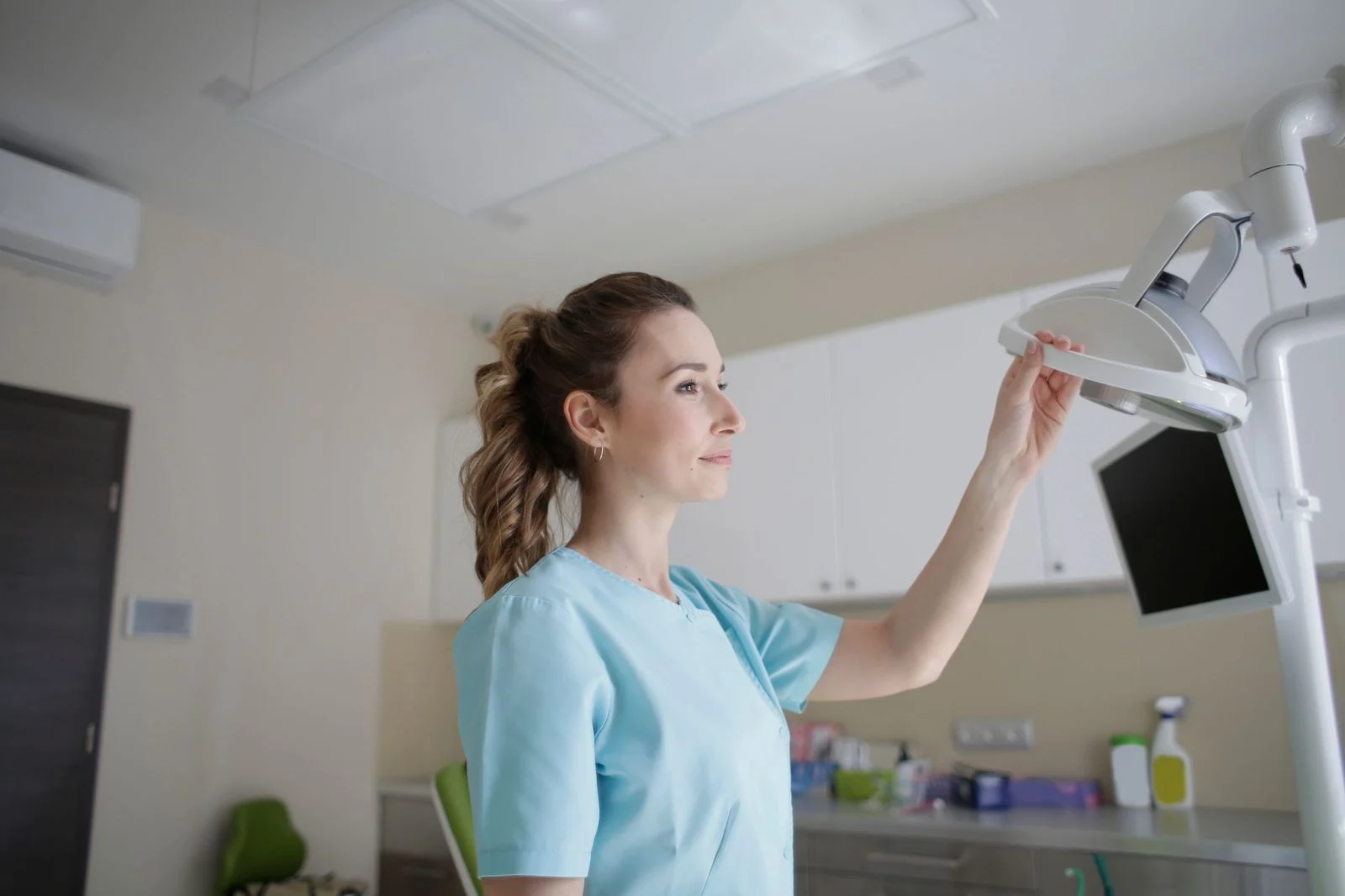 Mujer con uniforme azul de pie junto a una pantalla de computadora en una clínica luminosa, tocando una lámpara médica.