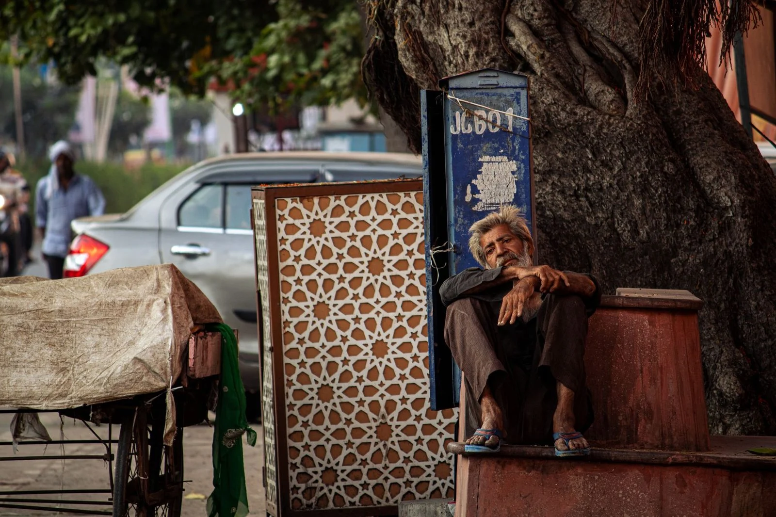 An elderly man rests against a tree on a busy Indian street, observing everyday city life.