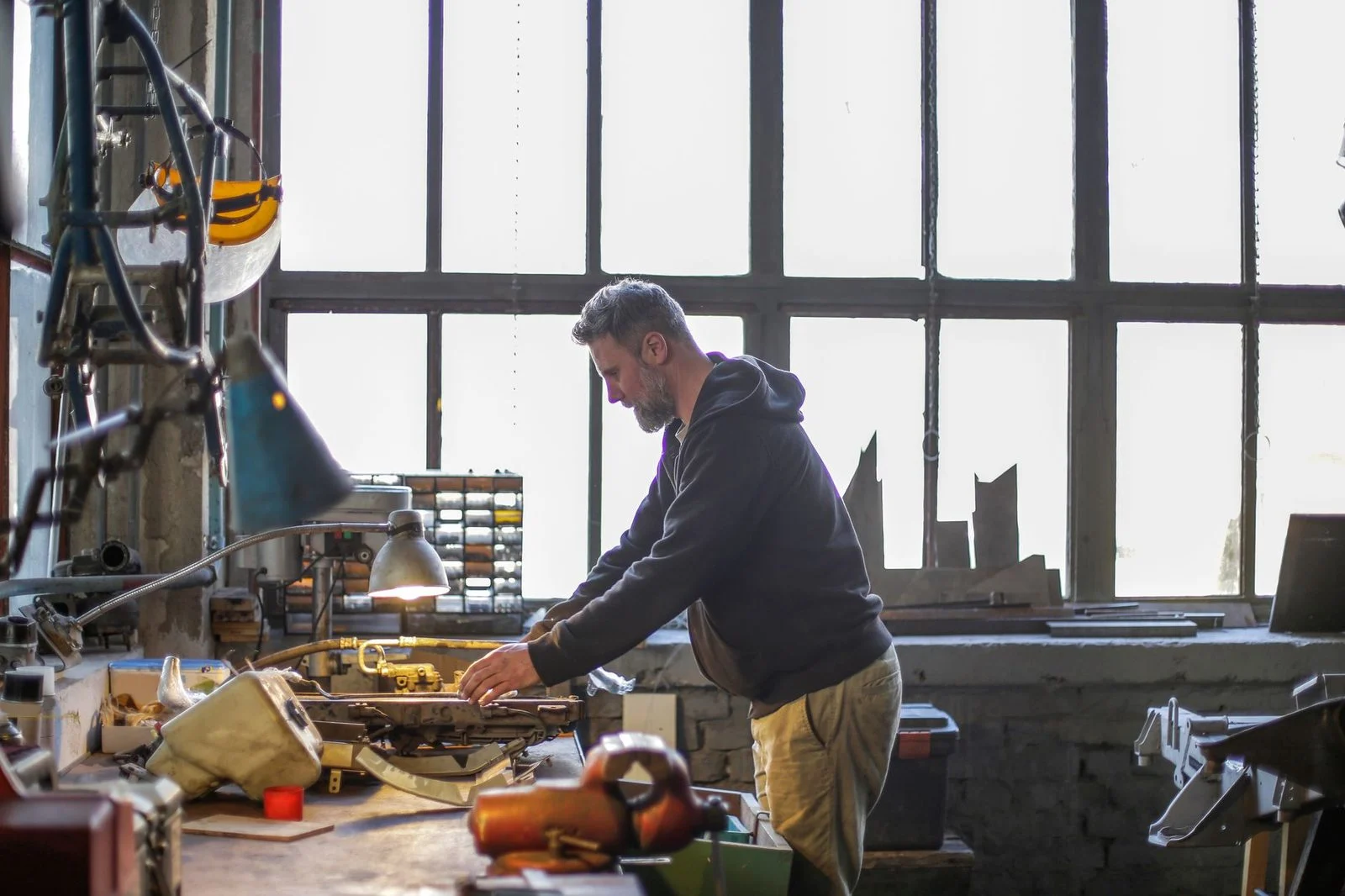 A bearded man in workwear fixes a broken detail with tools at a workbench in a workshop.