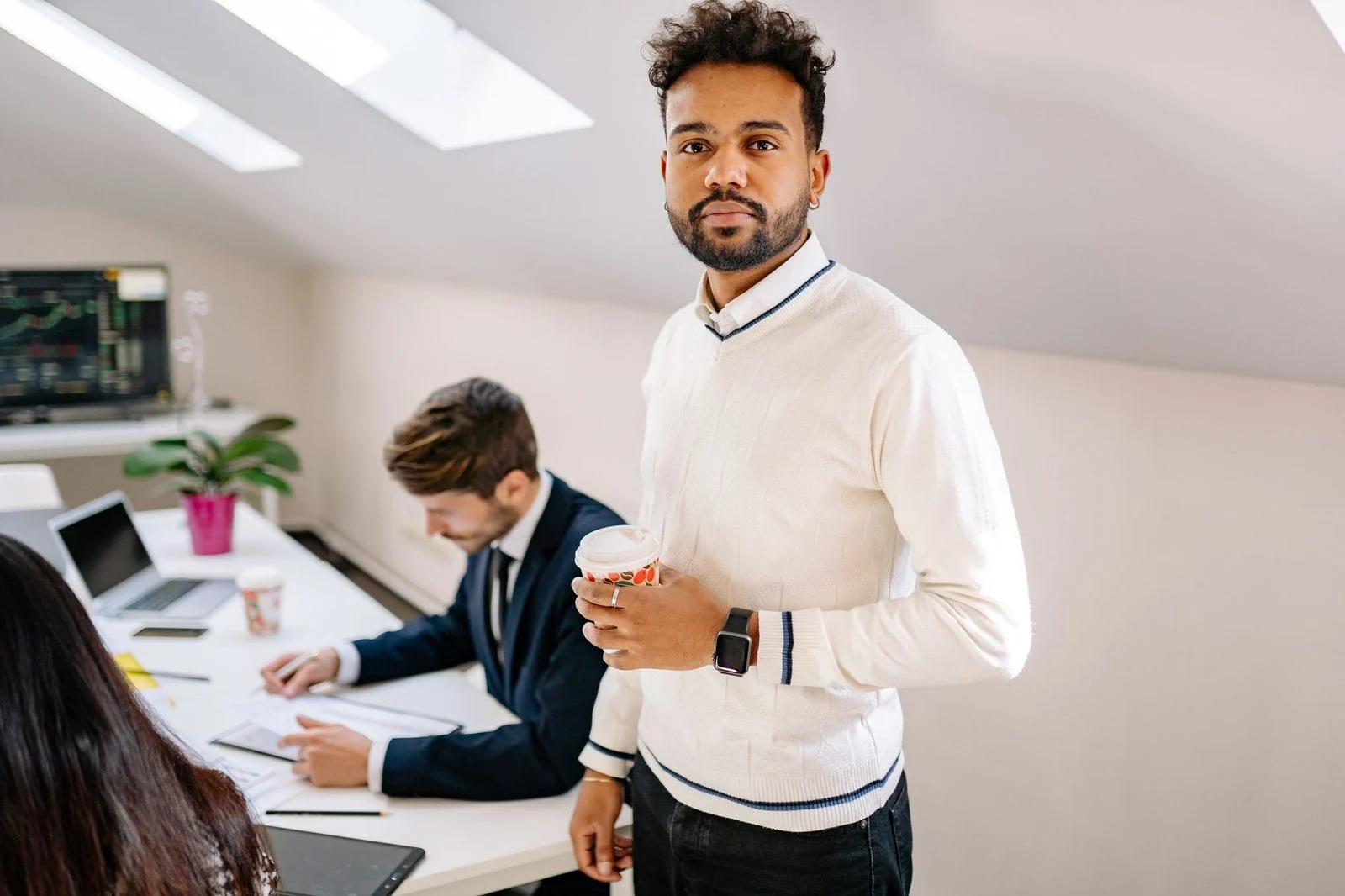 Businessman holding a coffee cup smiles confidently in a modern office while colleagues work nearby.