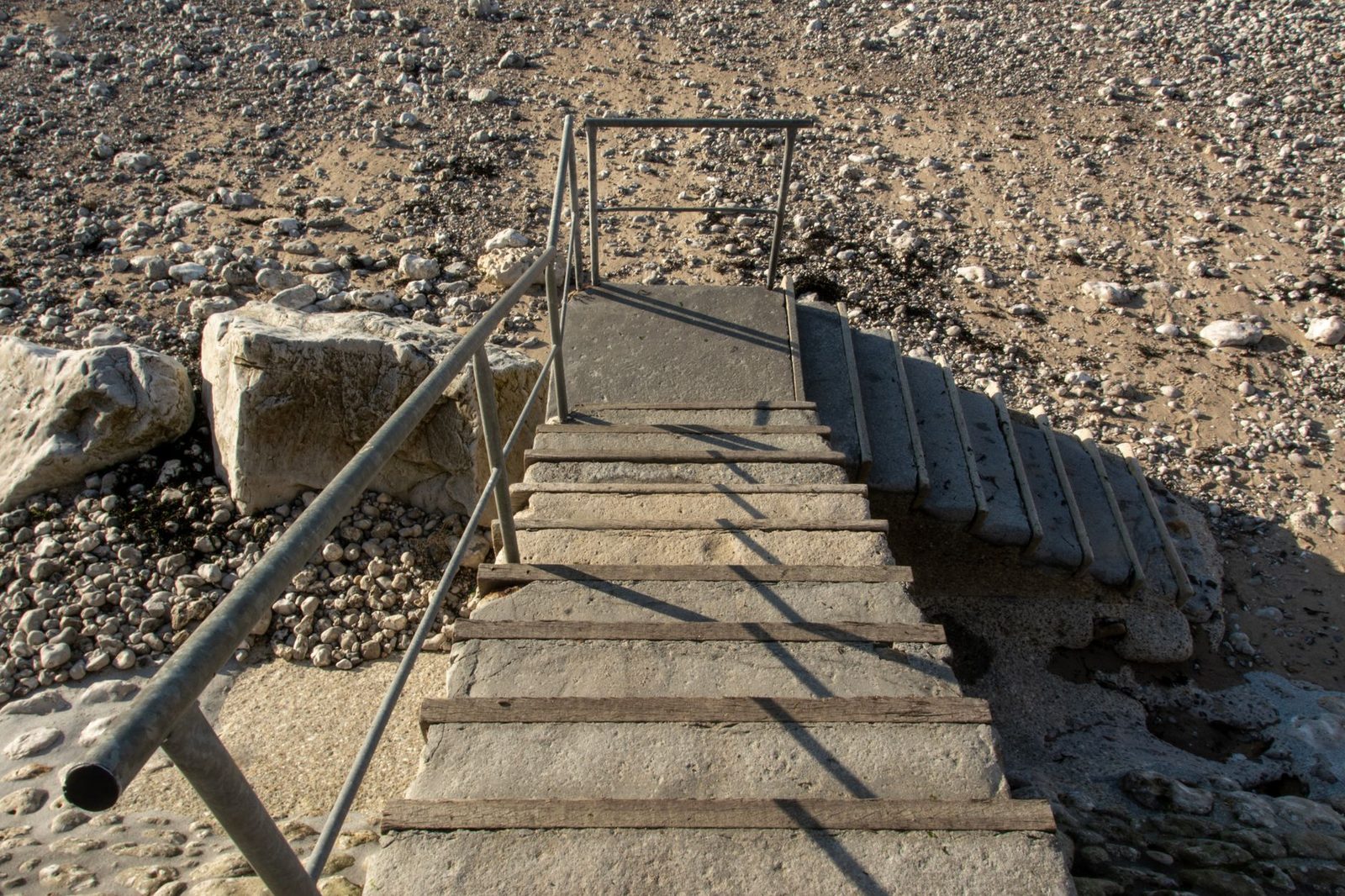Escaleras de hormigón descienden hacia una playa rocosa, proyectando sombras bajo el sol intenso.