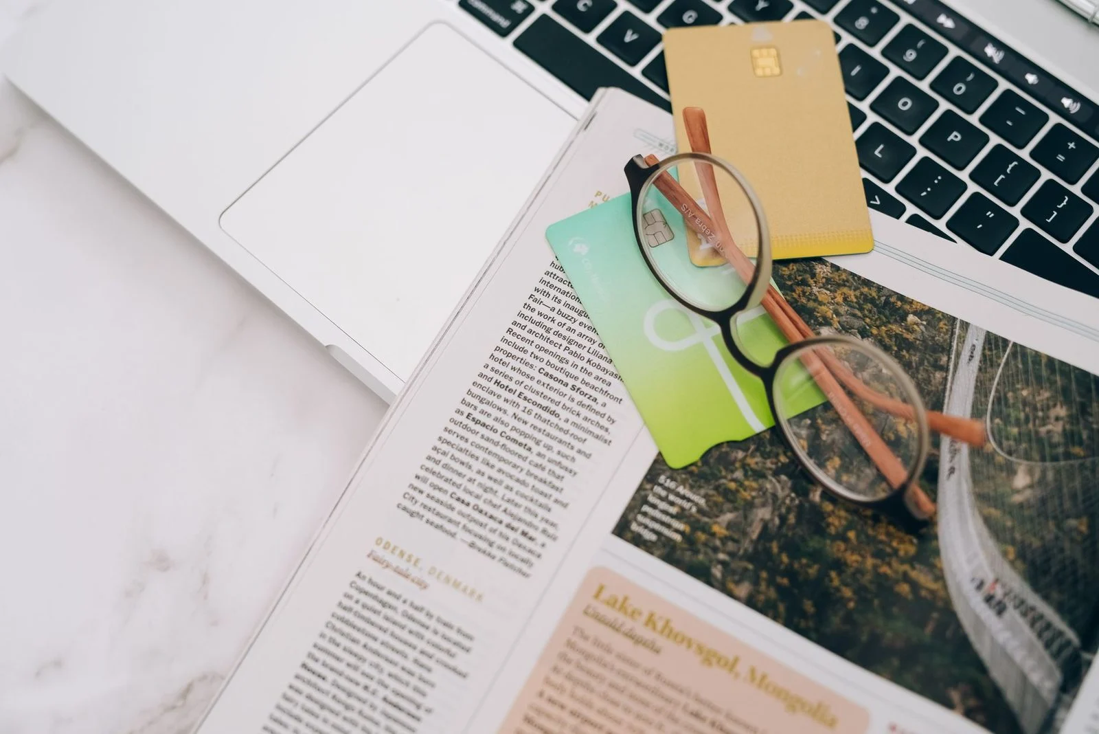 Eyeglasses rest on a magazine beside a laptop and credit cards on a table indoors.
