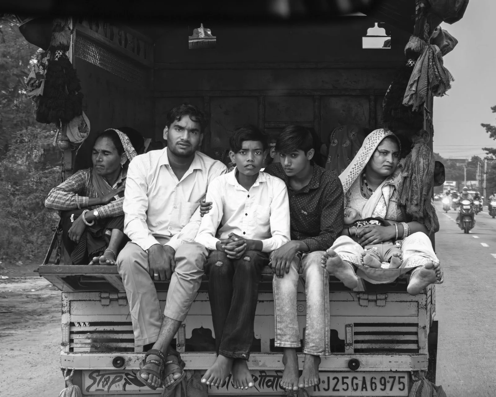 Family riding on the back of a vehicle along a rural road in India, shown in black and white.