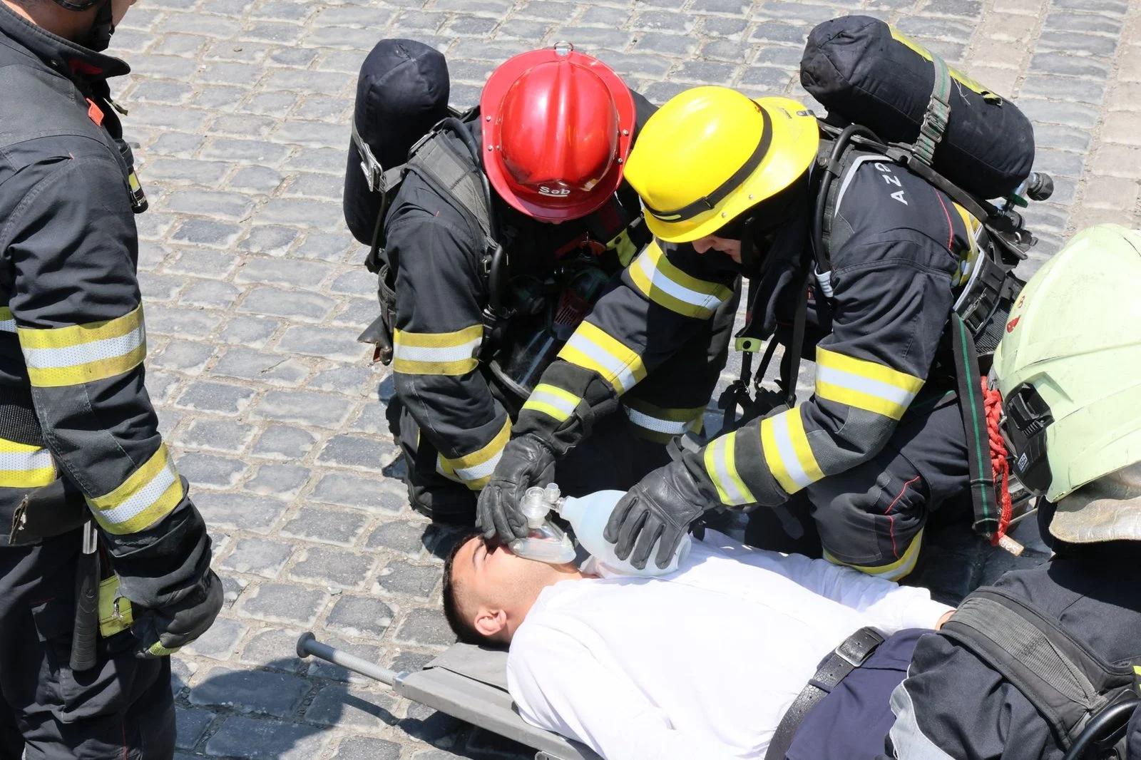 Bomberos realizando reanimación cardiopulmonar a un hombre herido con mascarilla de bolsa-válvula en una calle empedrada.