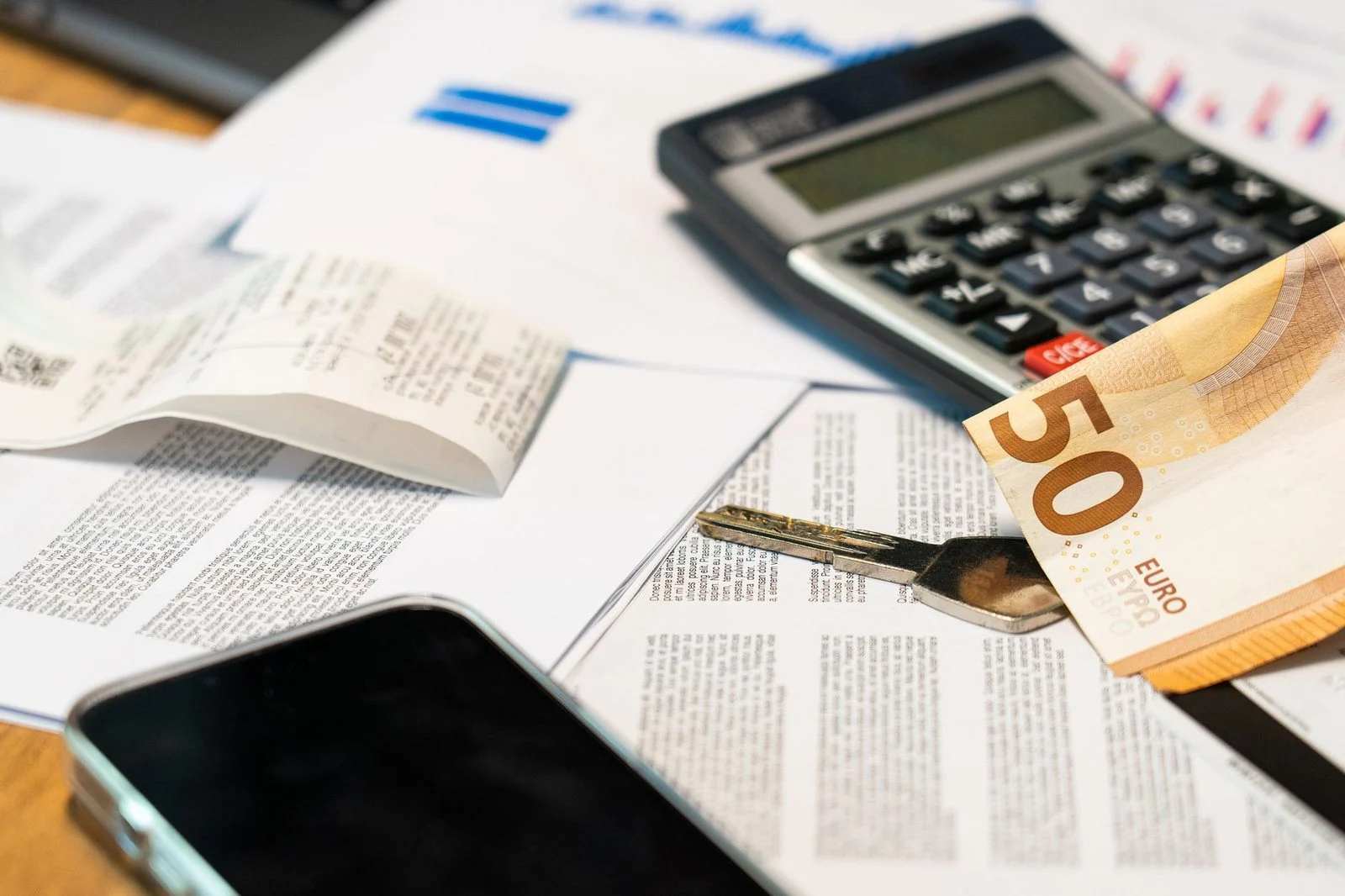 Documentos financieros, billetes de euro, una calculadora, un teléfono inteligente y una llave sobre un escritorio.