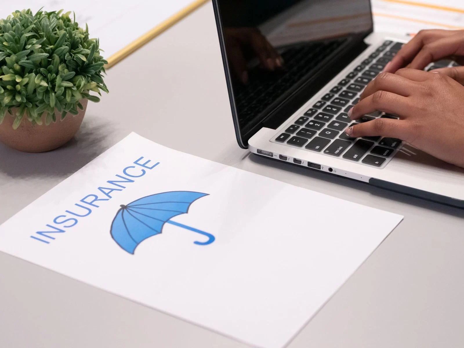 Hands typing on a laptop beside an insurance document on a desk.