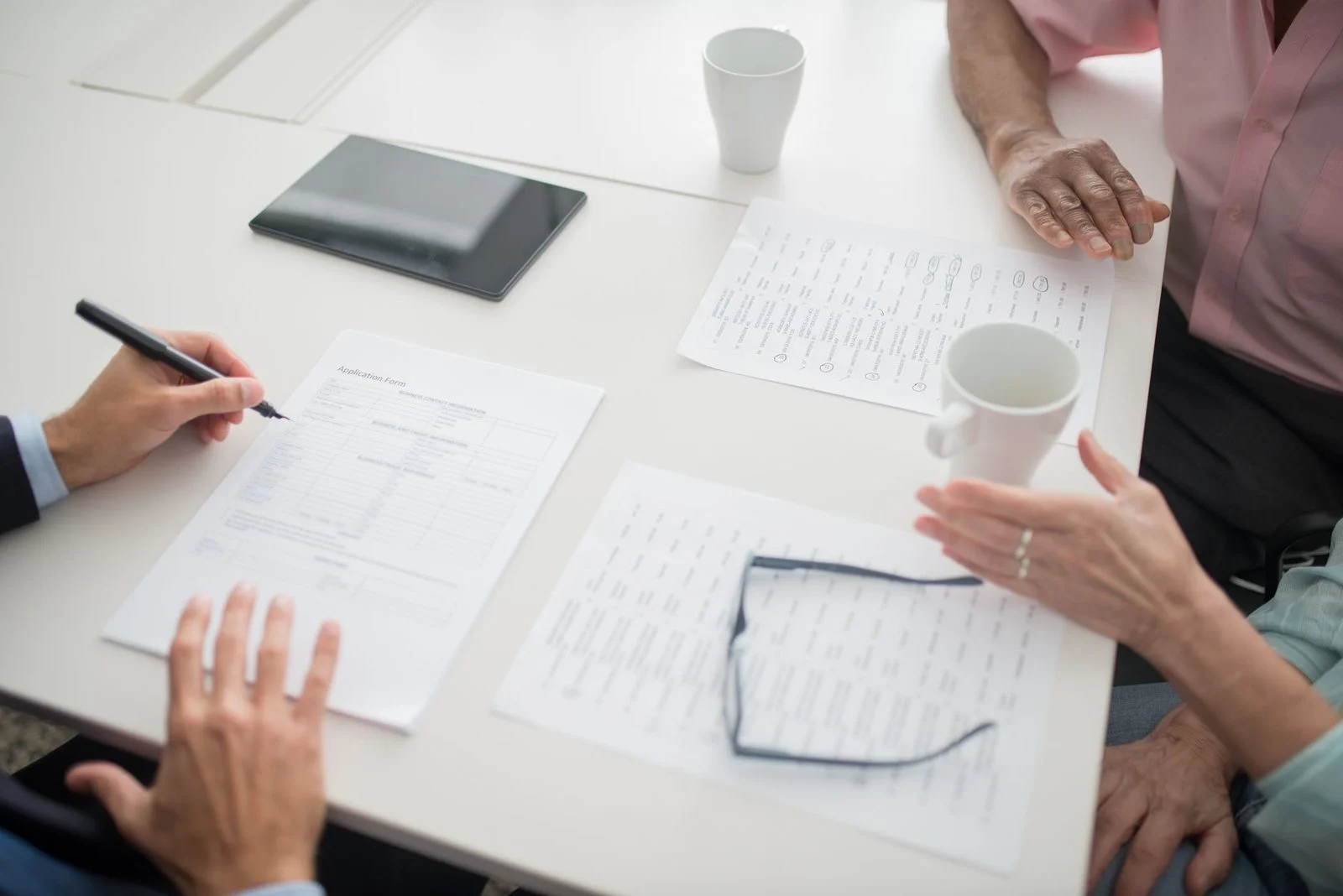 Hands writing on paperwork at a business meeting table with coffee cups and a tablet.