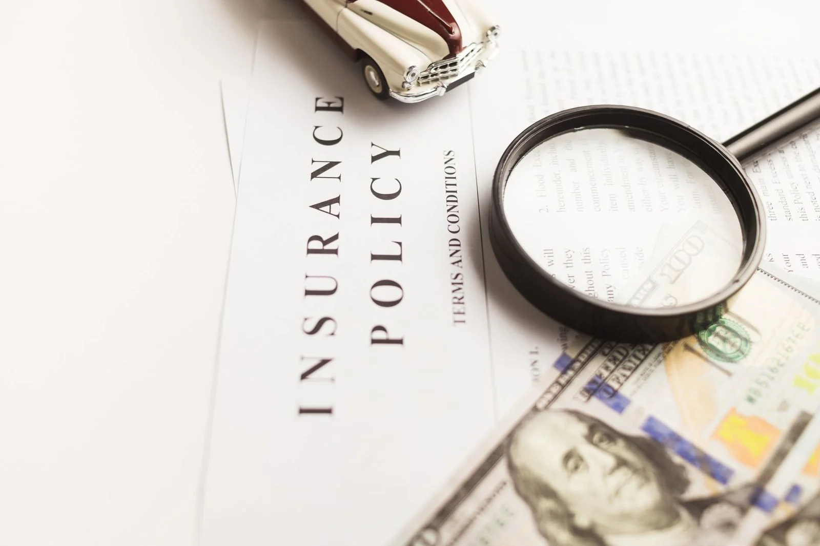 Close-up of an insurance policy document with a magnifying glass, cash, and a toy car on a table.