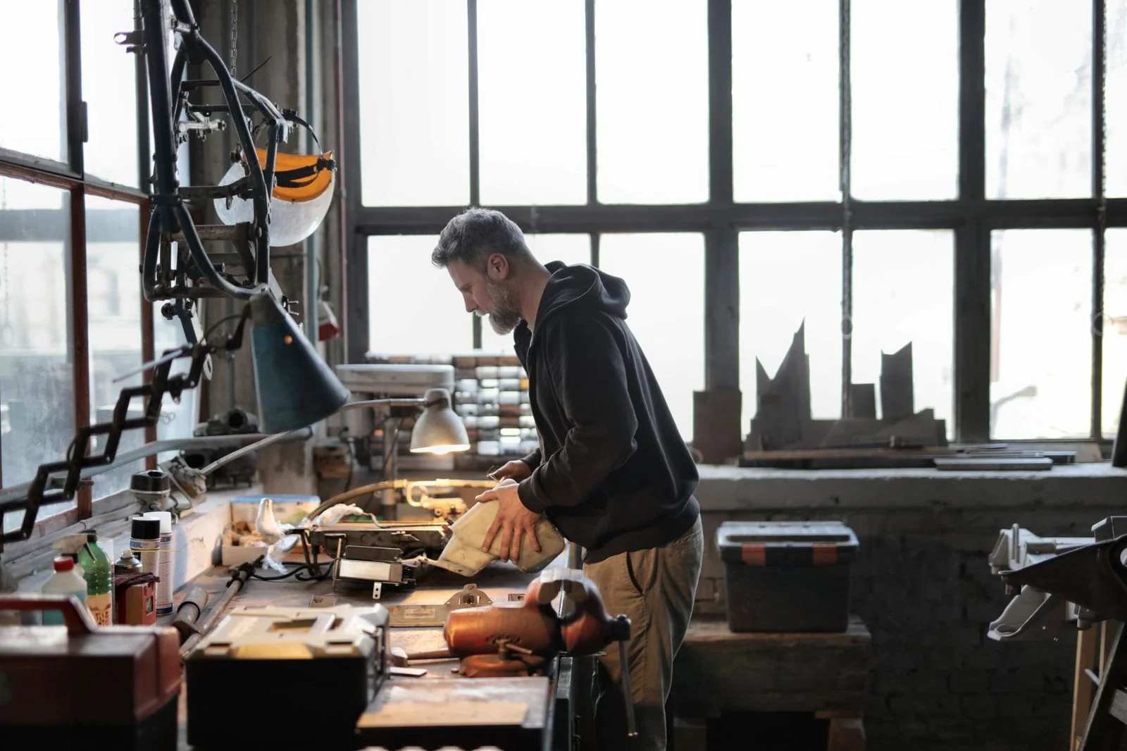 A male workshop owner stands beside a cluttered workbench, working on metal parts with tools.