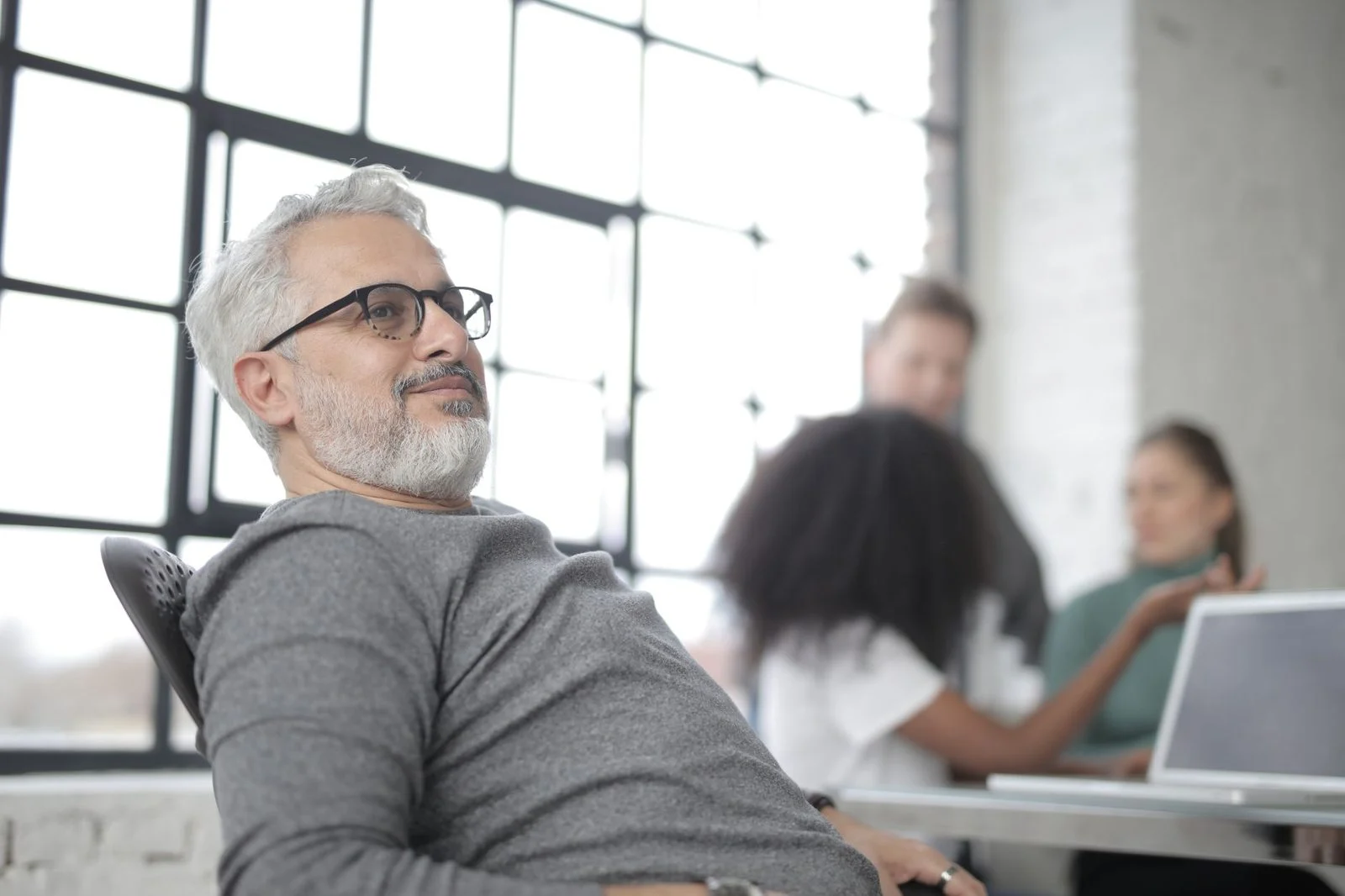 Older man with glasses leans back in an office chair at a desk with a laptop, while coworkers collaborate in an industrial-style workspace.