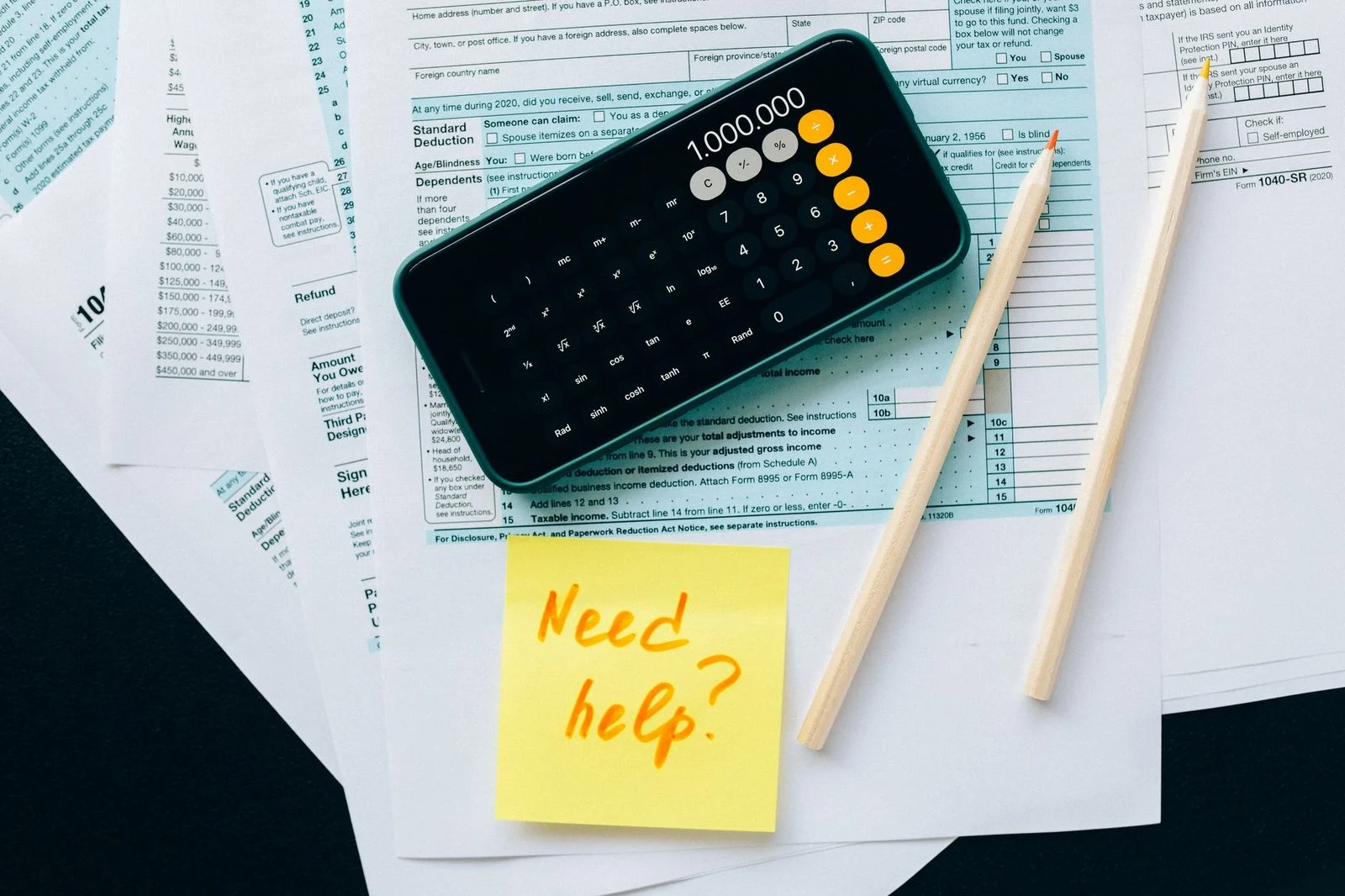 Vista superior de una calculadora, lápices y papeles financieros con una nota adhesiva que dice «¿Necesitas ayuda?».