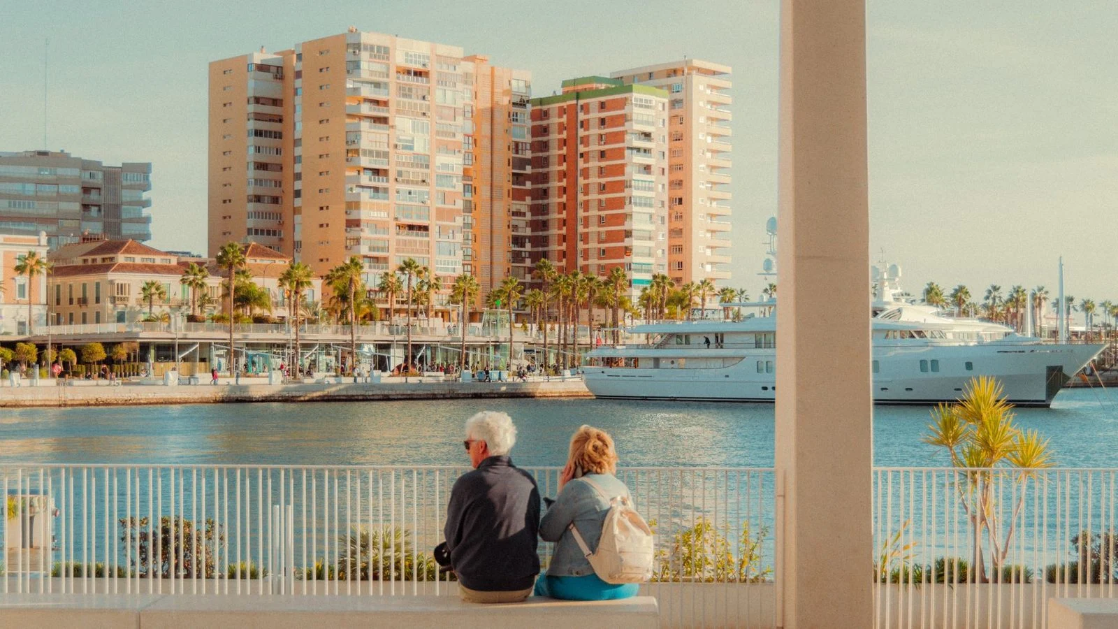 Pareja mayor disfrutando la vista en la marina de Málaga con edificios urbanos de fondo.