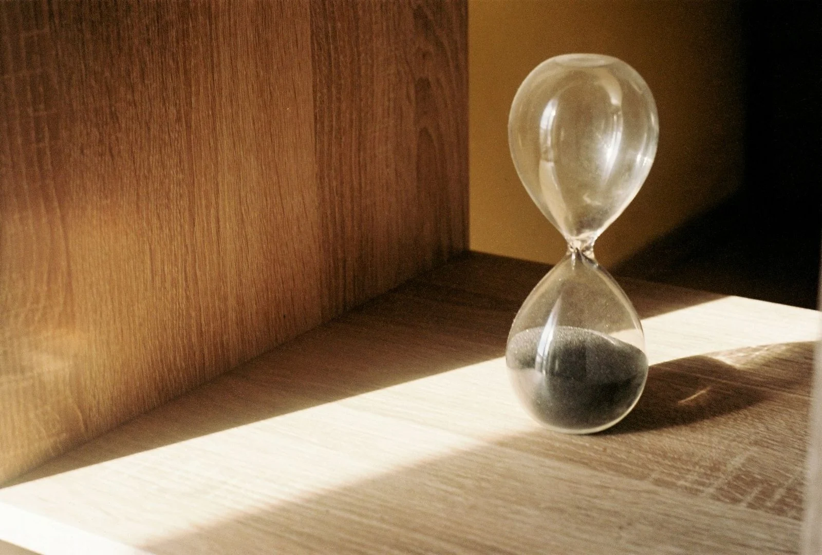Reloj de arena de vidrio con arena negra sobre una repisa de madera iluminada por luz natural.