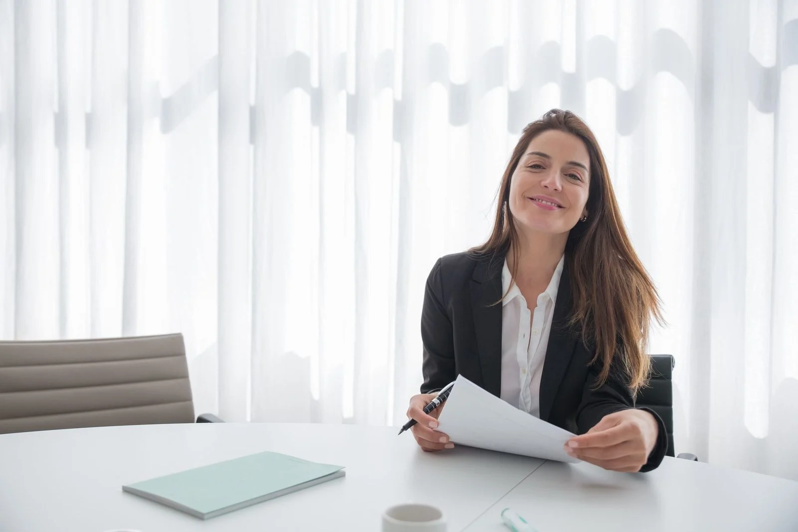 Smiling businesswoman in a blazer holding a paper at an indoor office table.