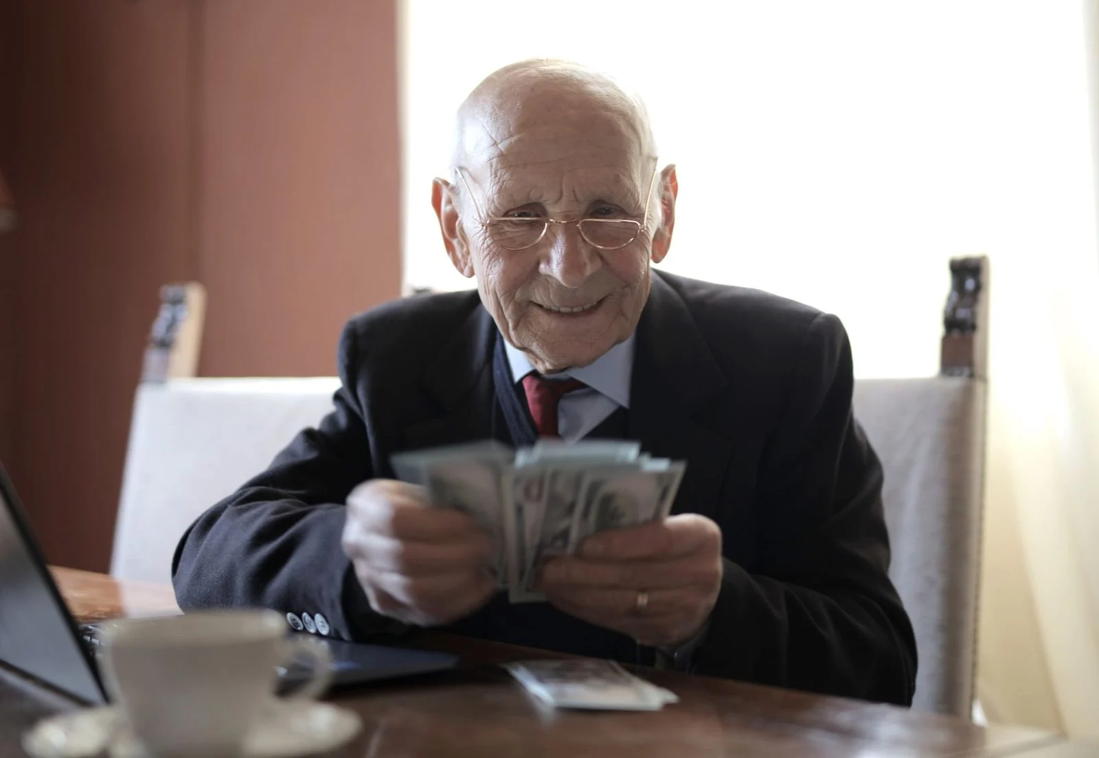 Empresario mayor sonriente con gafas cuenta dinero en un escritorio de madera junto a una laptop y una taza de café.