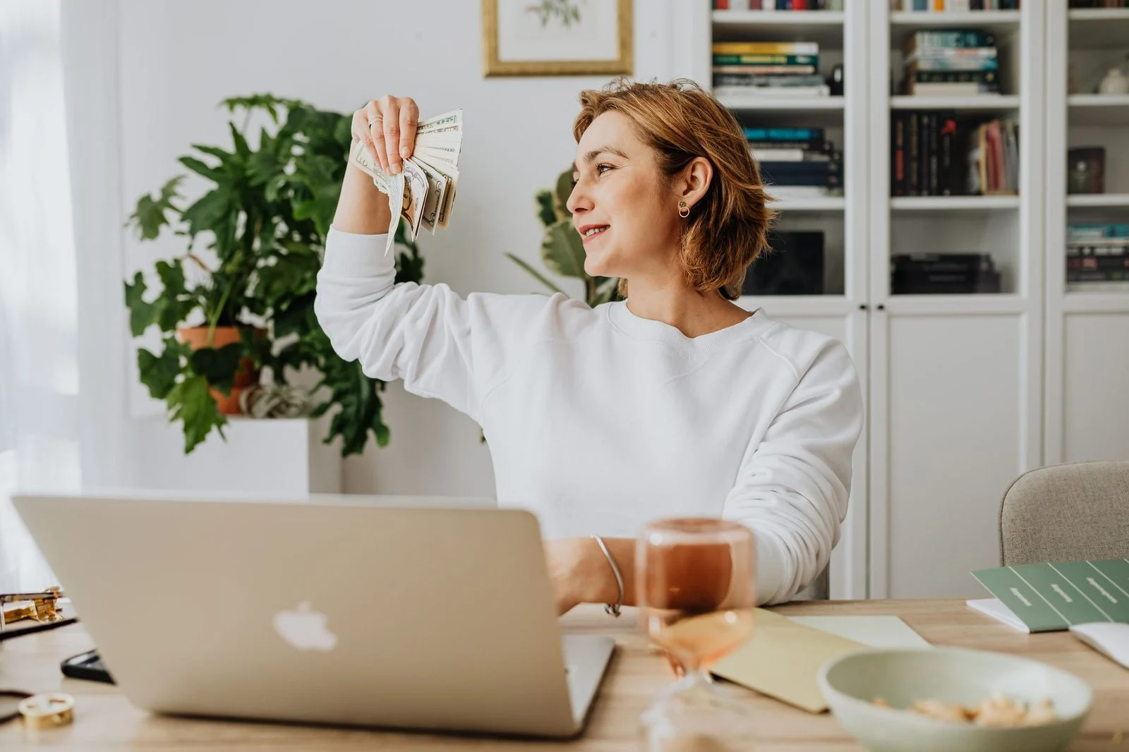 Mujer sonriente sosteniendo billetes mientras trabaja en una laptop en un escritorio de oficina en casa.