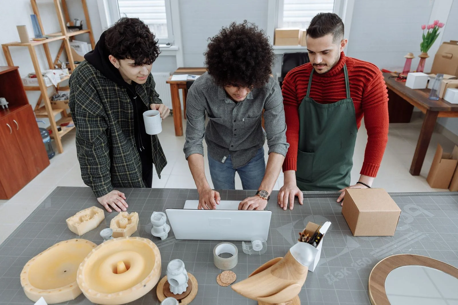 Tres hombres trabajan juntos en una mesa de taller con una computadora portátil y materiales de manualidades.