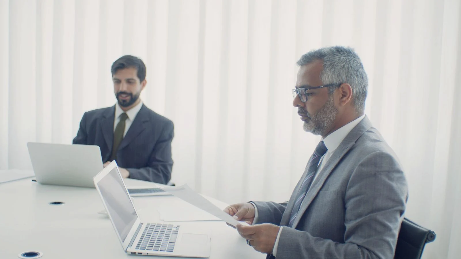 Two colleagues sit at a desk in an office, reviewing documents and working on laptops together.