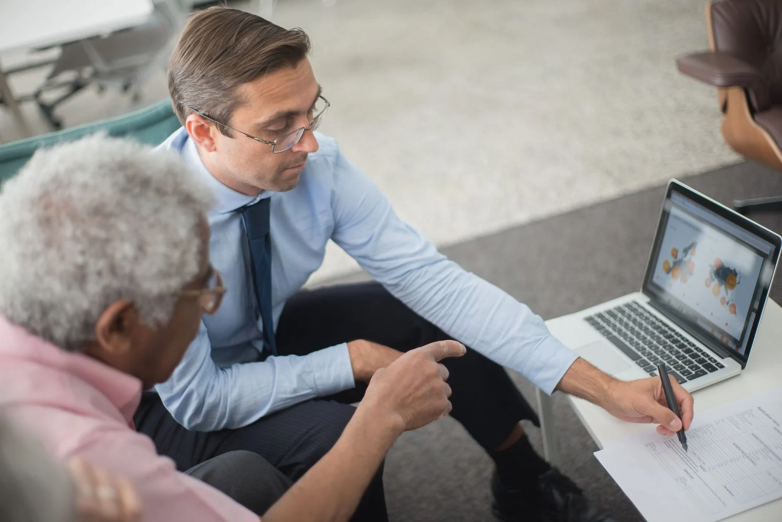 Two men in a business meeting review financial documents and discuss data on a laptop.