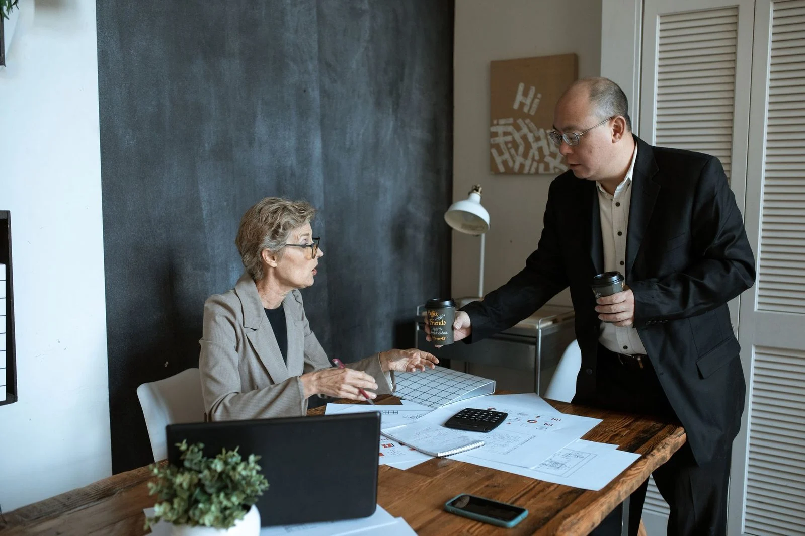 Two professionals chat over coffee at a modern office desk beside papers and a laptop.