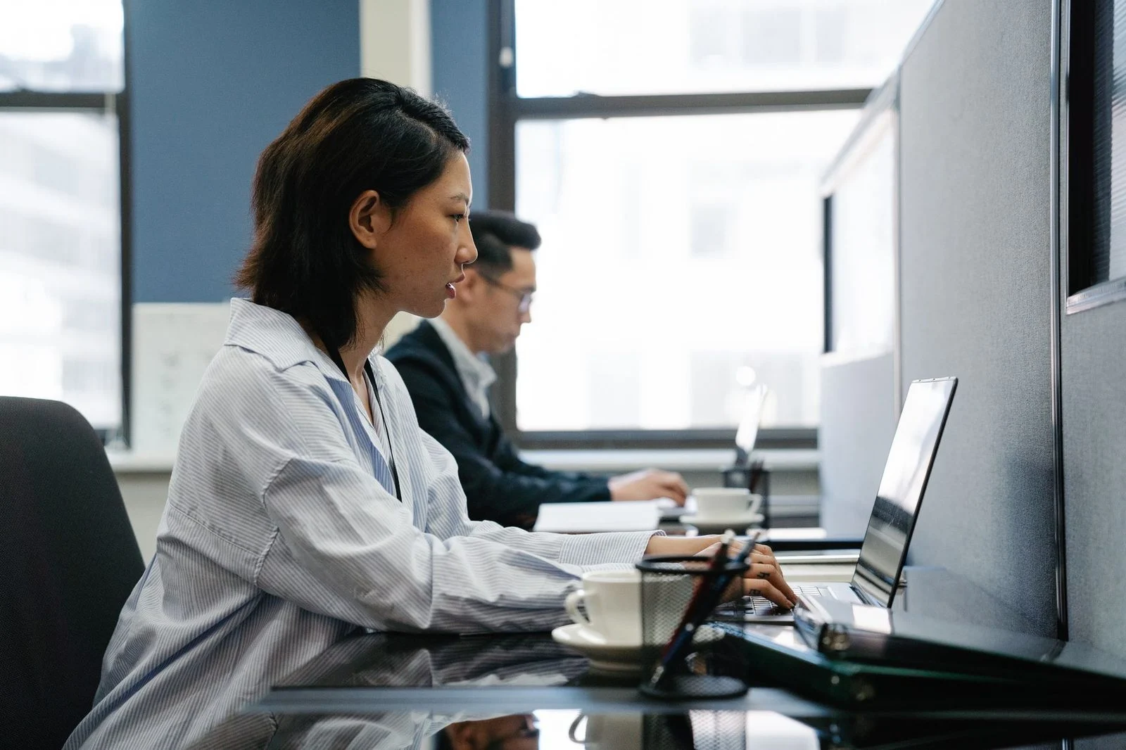 Two professionals work side by side on laptops in a modern office, collaborating on a shared task.