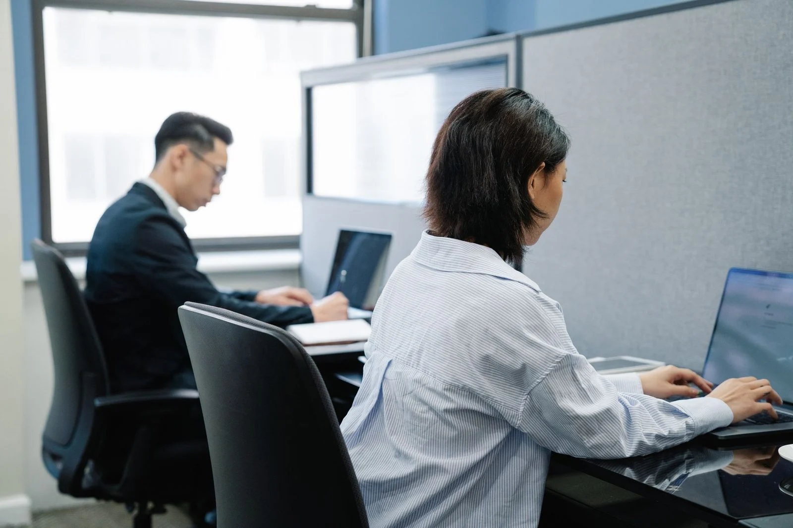 Two professionals work on laptops at adjacent desks in a modern office.
