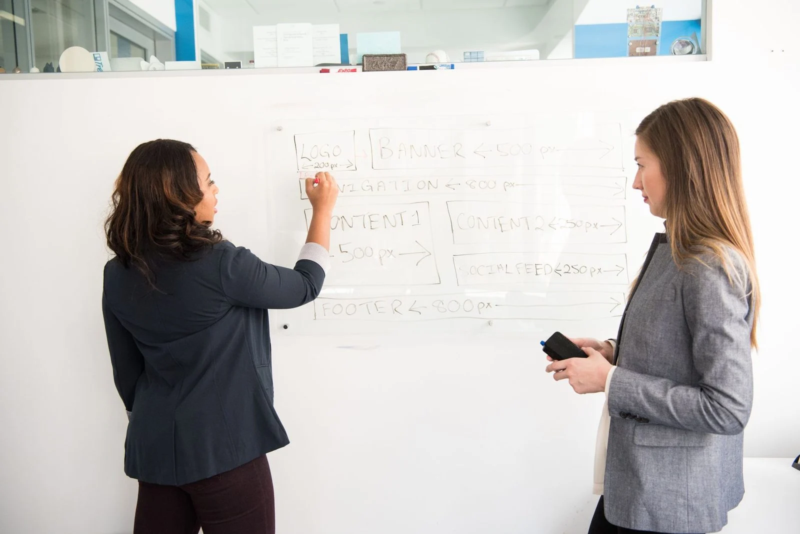 Two women stand at a whiteboard in an office, discussing a presentation.