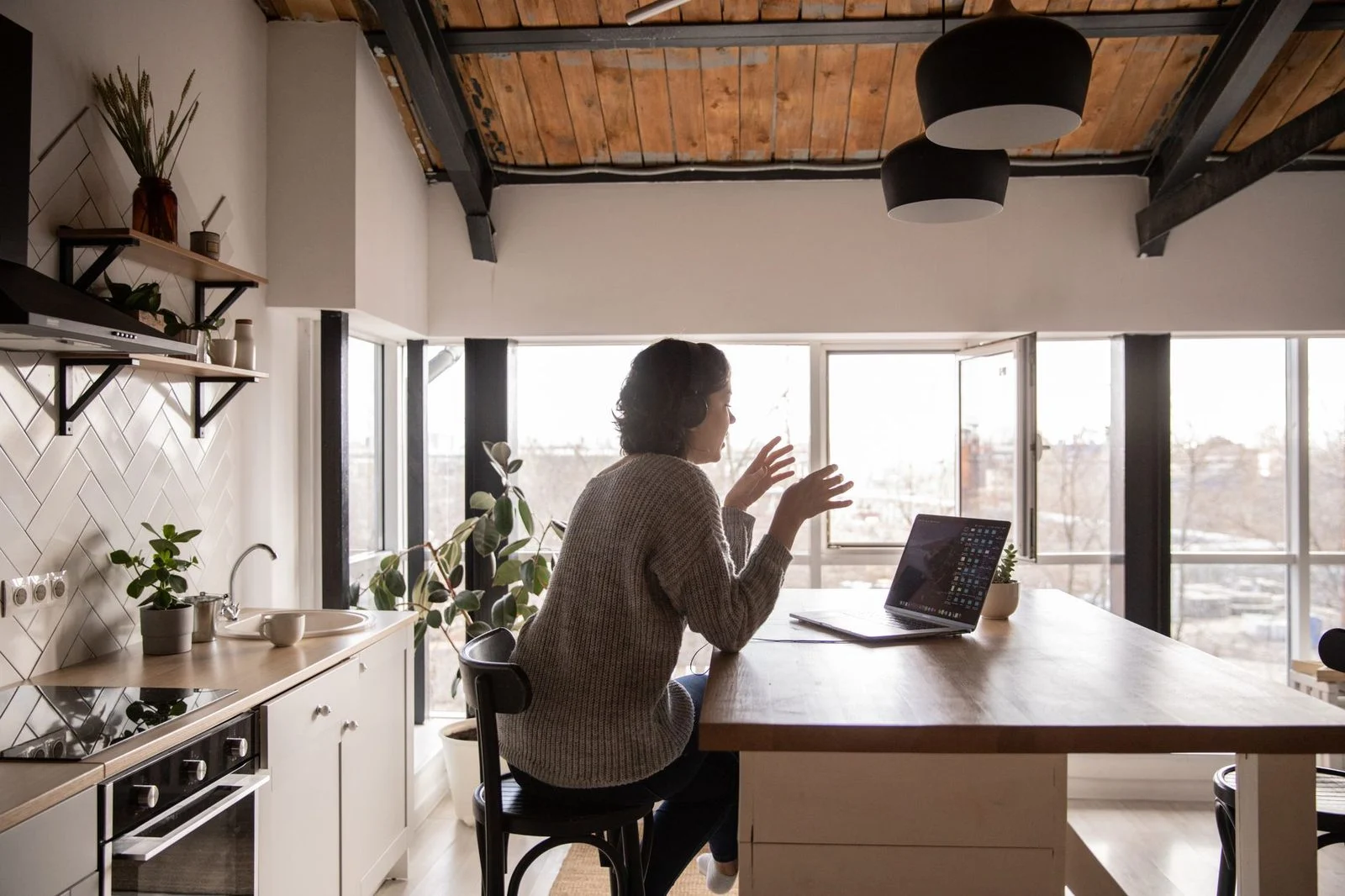 Young woman seated at a wooden kitchen table, turned away from the camera, talking expressively during a video call on a laptop.